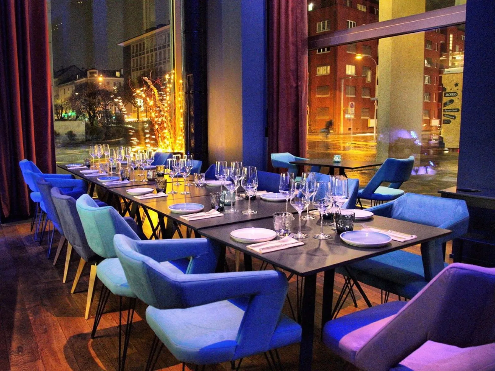 Ein stilvoll gedeckter Tisch in einem modernen Restaurant mit bunten Stühlen und großen Fenstern, die einen nächtlichen Blick auf eine beleuchtete Stadtstraße bieten.