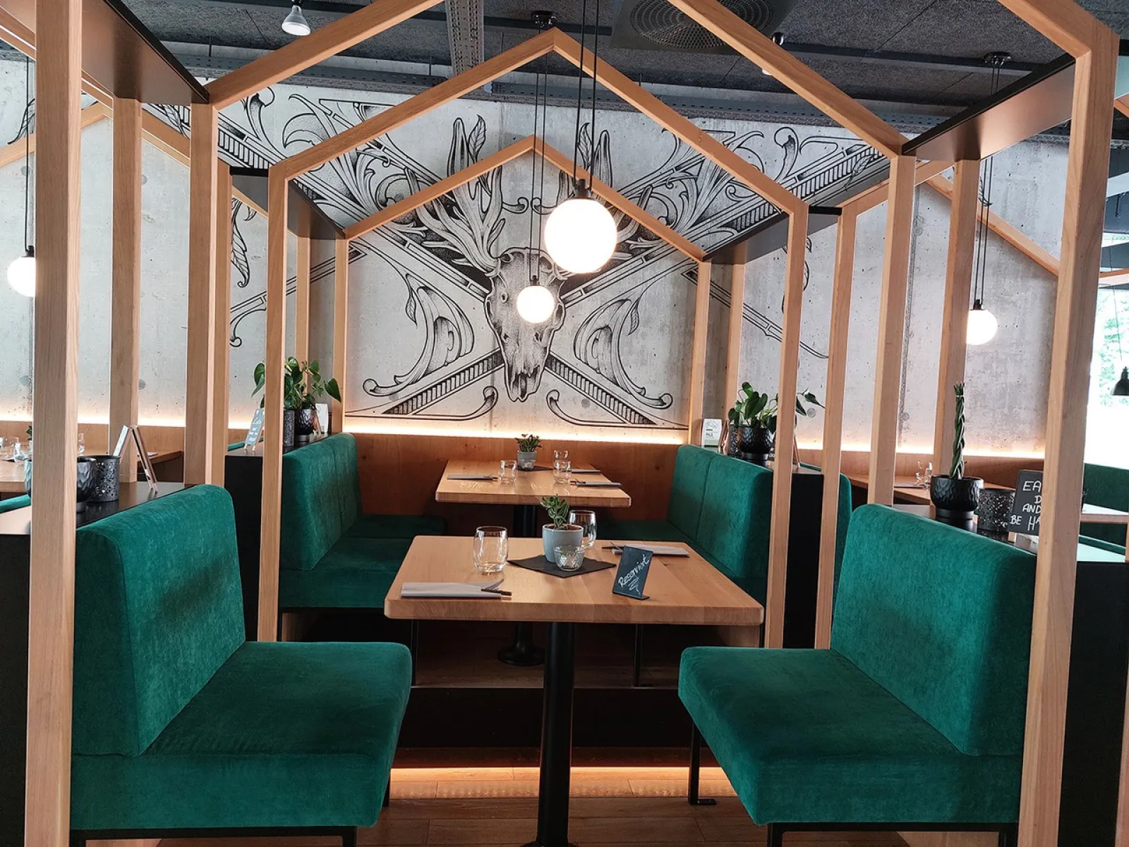 Ein stilvoll eingerichteter Restaurantbereich mit grünen Polsterbänken und Holztischen, umgeben von einem hölzernen Rahmen. Im Hintergrund ist eine kunstvolle Wandzeichnung eines Hirschkopfes zu sehen. Dezente Beleuchtung schafft eine einladende Atmosphäre.