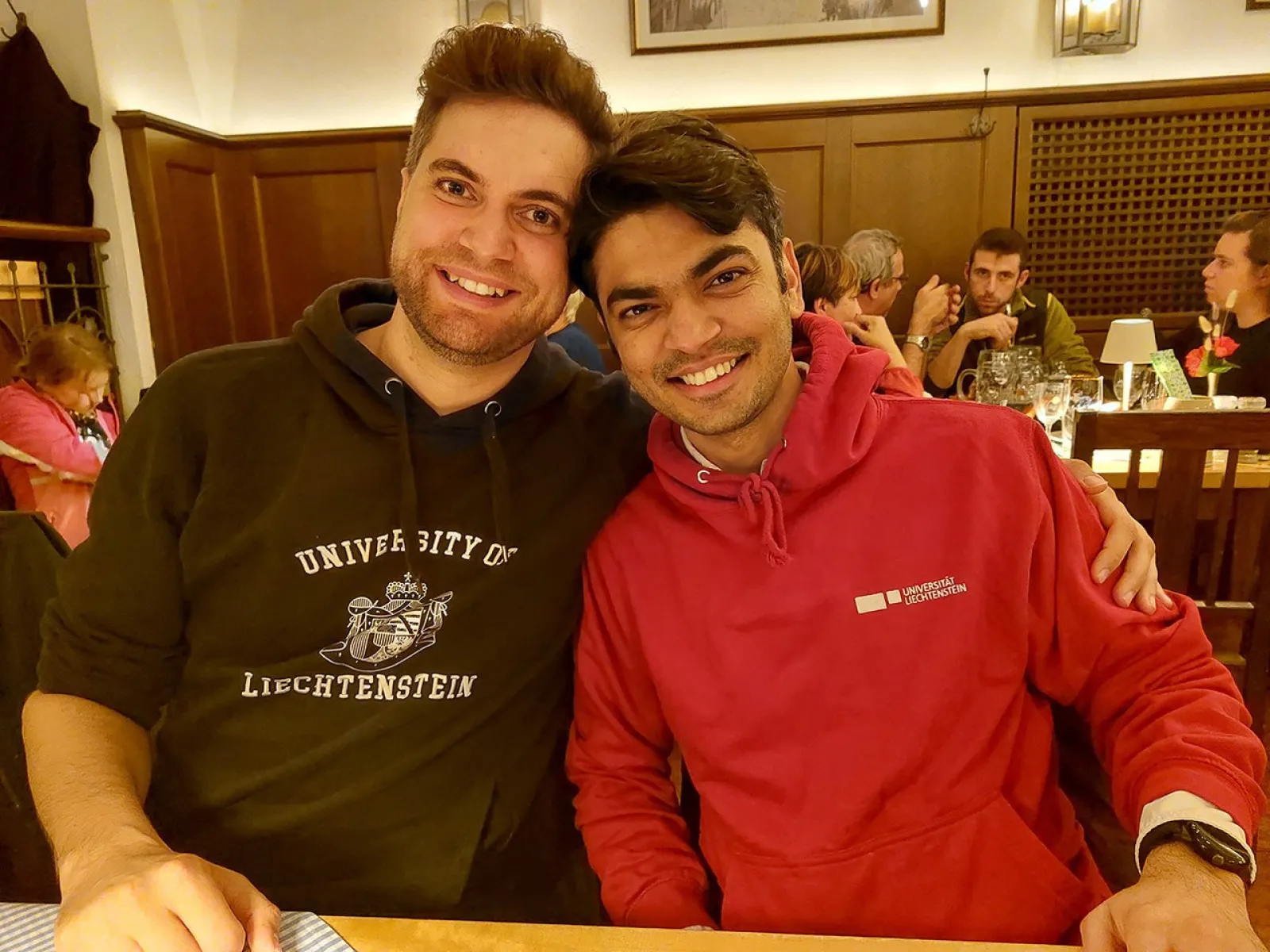 Zwei Personen sitzen lächelnd in einem Restaurant. Beide tragen Kapuzenpullover, einer mit dem Logo der Universität Liechtenstein. Im Hintergrund sind weitere Gäste an Tischen zu sehen. Die Atmosphäre wirkt gemütlich und gesellig.