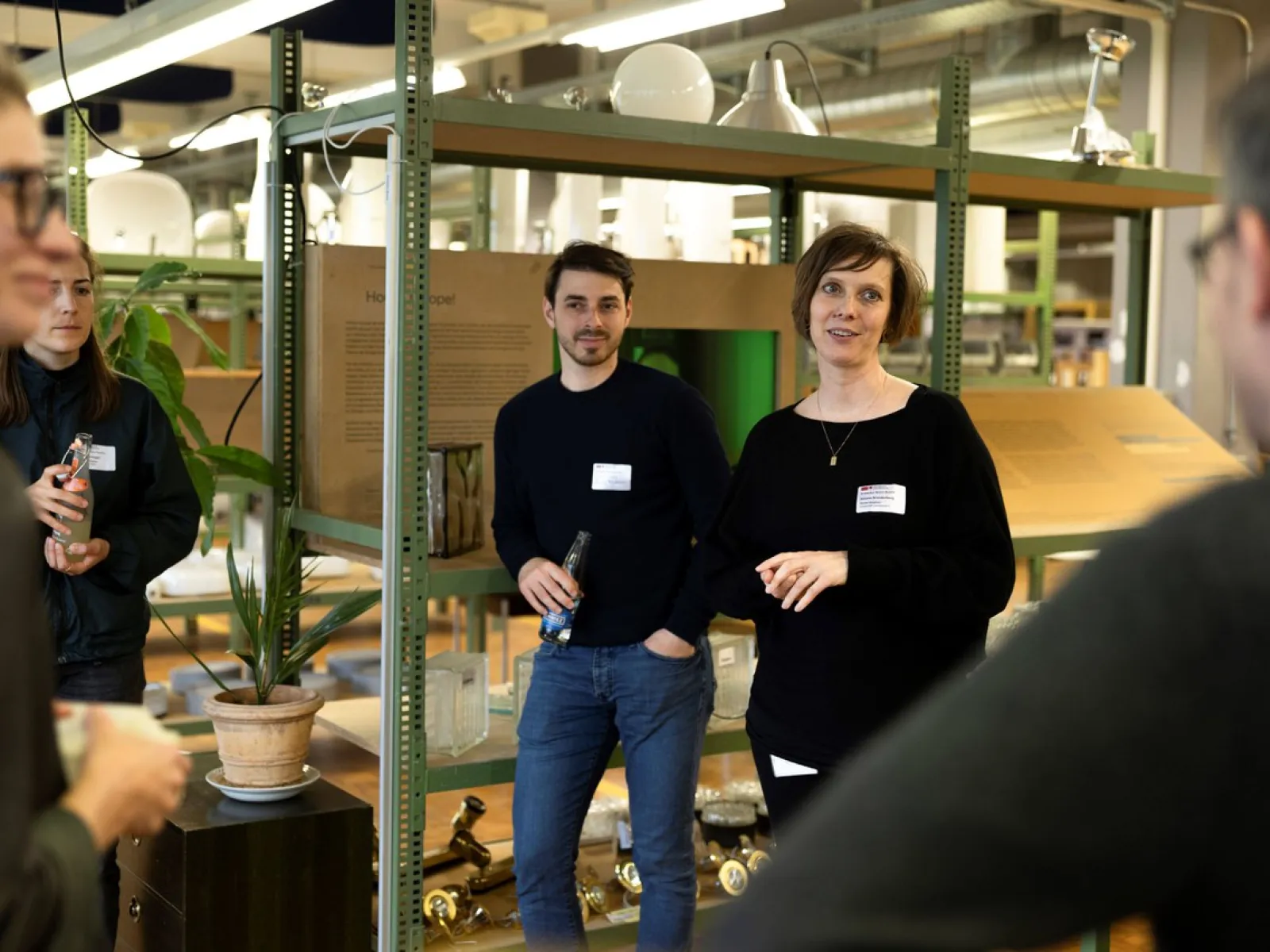 Eine Gruppe von Menschen steht in einem Innenraum vor Regalen mit Baumaterialien. Zwei Personen tragen Namensschilder und lächeln in die Kamera. Im Hintergrund sind Pflanzen und Lampen zu sehen.