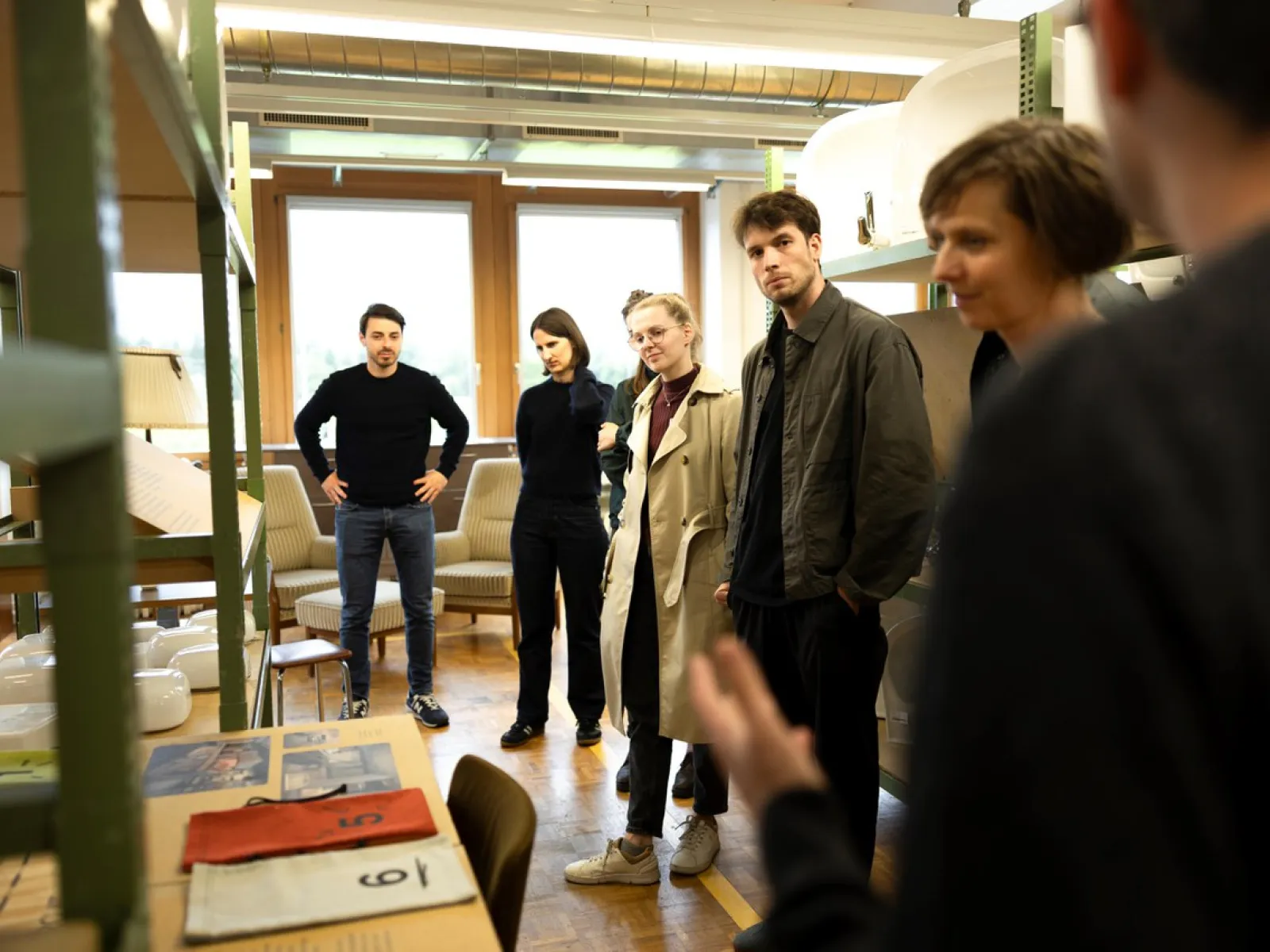 Eine Gruppe von Menschen steht in einem Raum mit Regalen und Möbeln. Sie hören einer Person zu, die außerhalb des Bildes spricht. Die Szene wirkt wie ein informelles Treffen oder eine Führung.