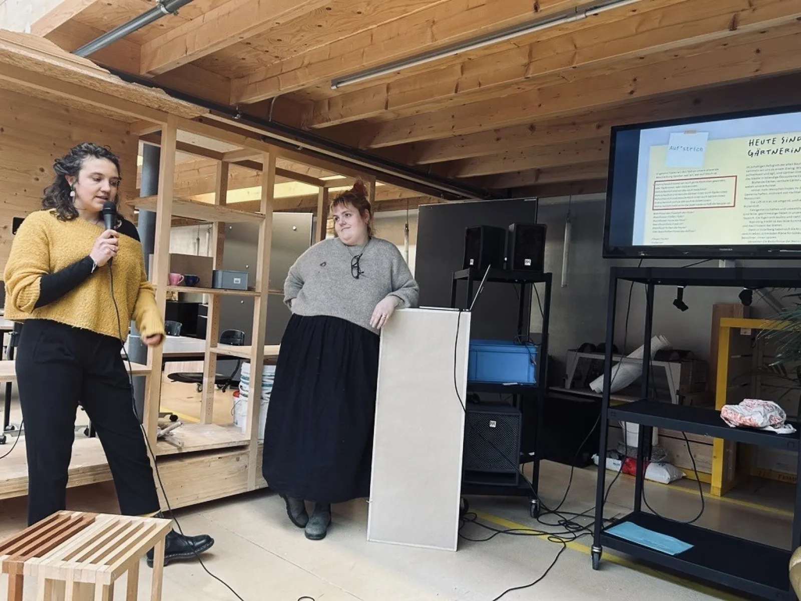 Zwei Personen präsentieren in einem Raum mit Holzelementen. Eine Person spricht in ein Mikrofon, die andere steht neben einem Rednerpult. Im Hintergrund ist eine Leinwand mit einer Präsentation zu sehen.