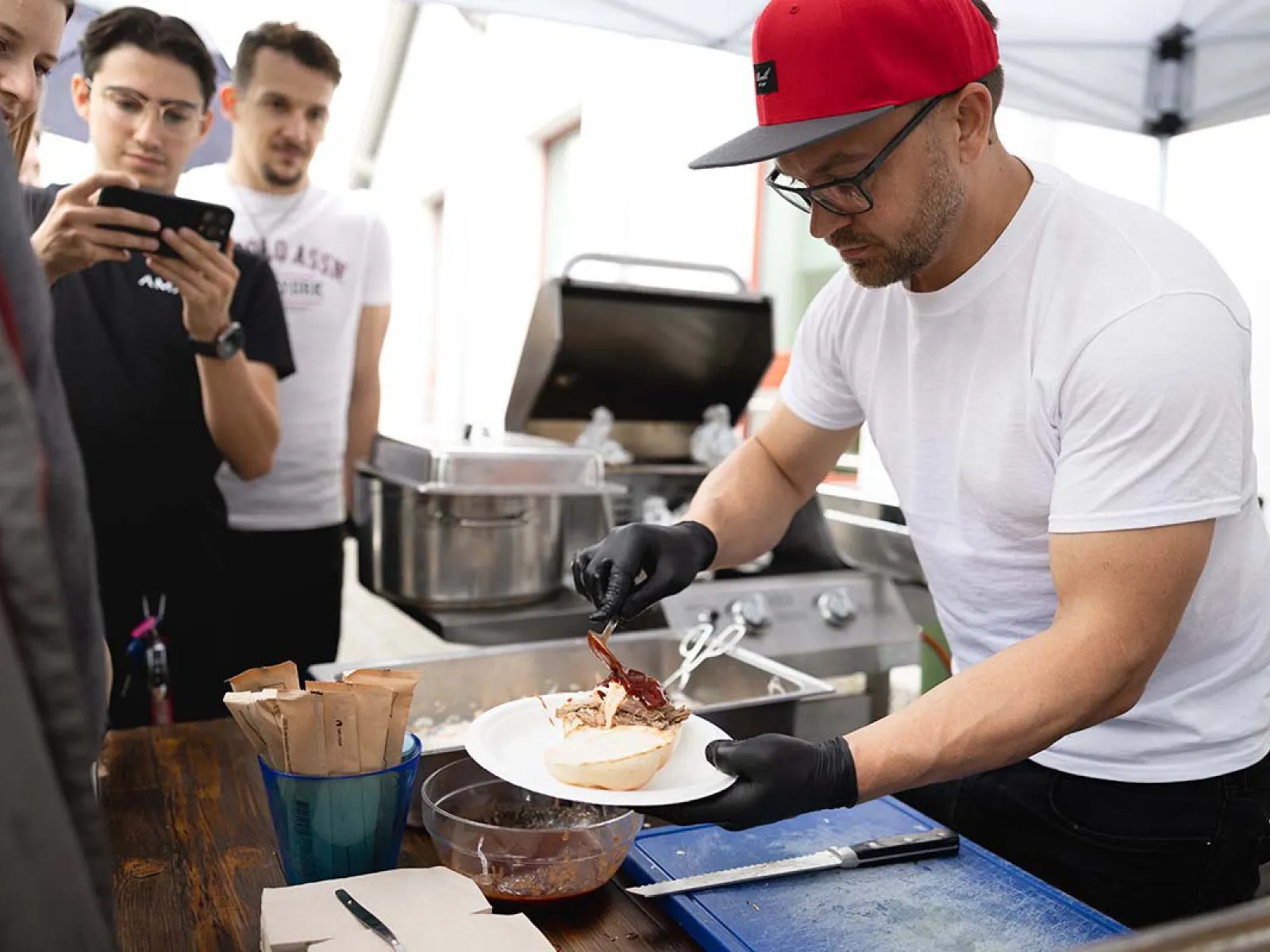 Ein Mann mit roter Kappe und schwarzen Handschuhen bereitet Essen auf einem Teller vor. Im Hintergrund stehen mehrere Personen und beobachten ihn. Eine Grillstation ist ebenfalls zu sehen. Die Szene findet im Freien unter einem Zelt statt.