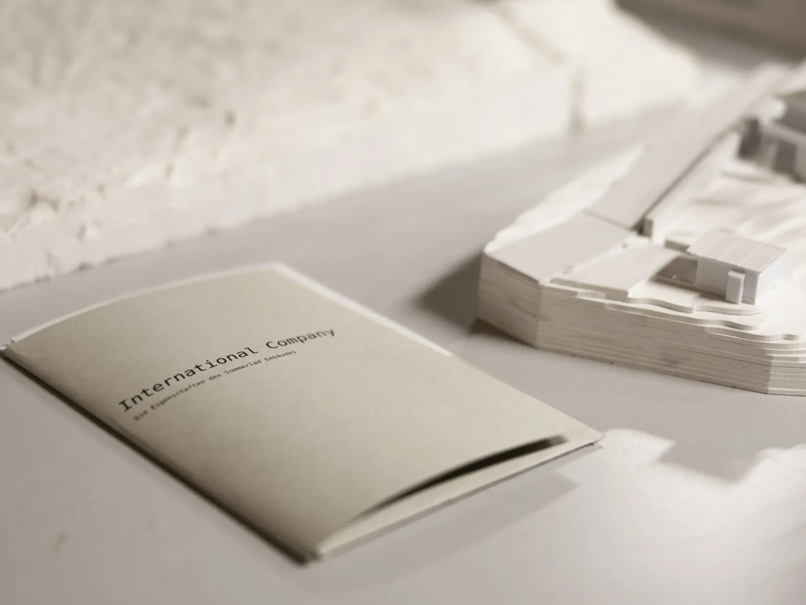 Ein Heft mit der Aufschrift "International Company" liegt auf einem Tisch neben einem Architekturmodell. Das Bild vermittelt eine professionelle und kreative Atmosphäre, passend für eine universitäre Umgebung.
