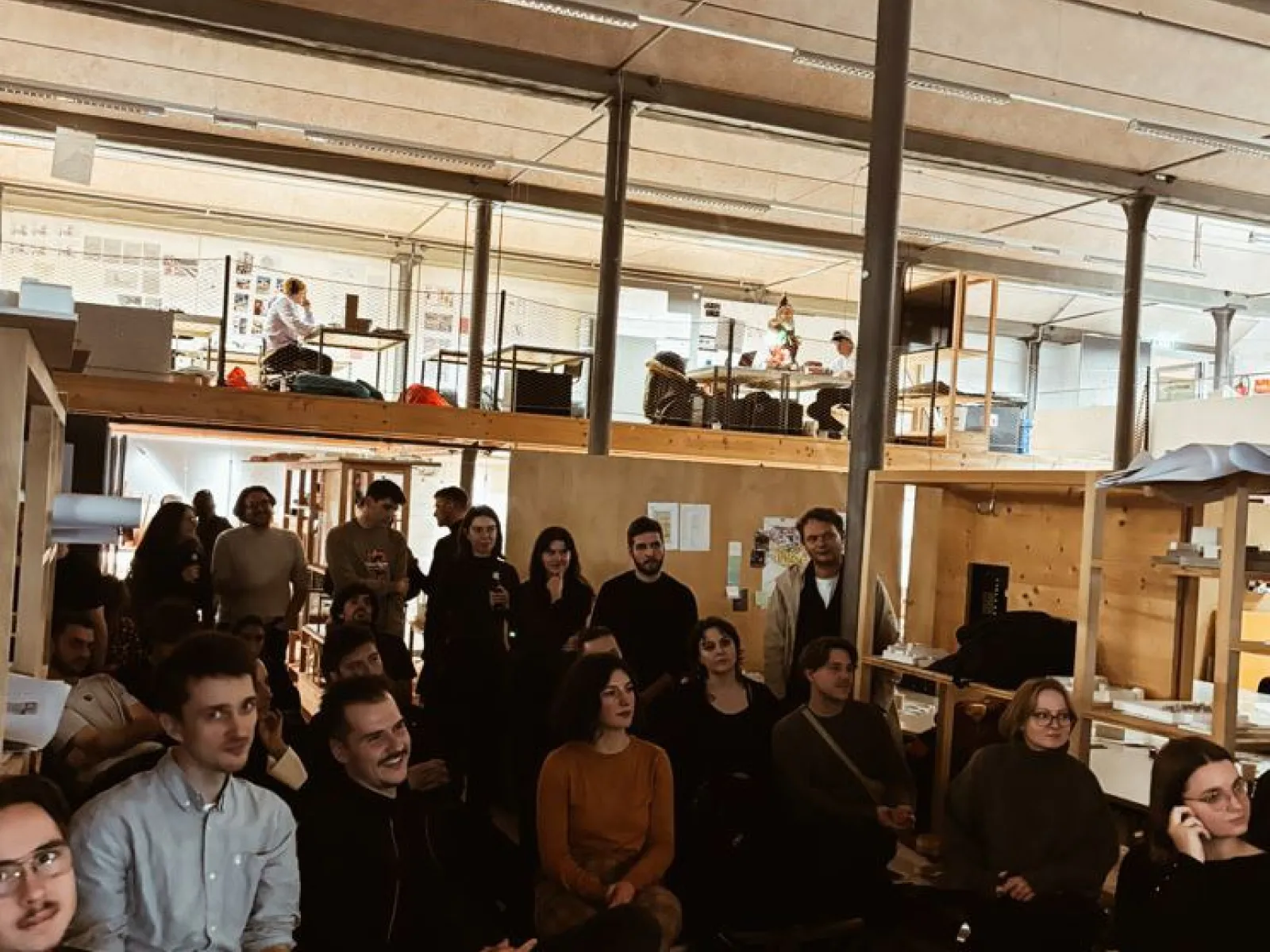 Eine Gruppe von Studierenden und Alumni sitzt in einem Architekturstudio und hört einer Präsentation zu. Im Hintergrund sind Arbeitsplätze und eine Uhr an der Wand zu sehen. Die Atmosphäre ist konzentriert und interessiert.