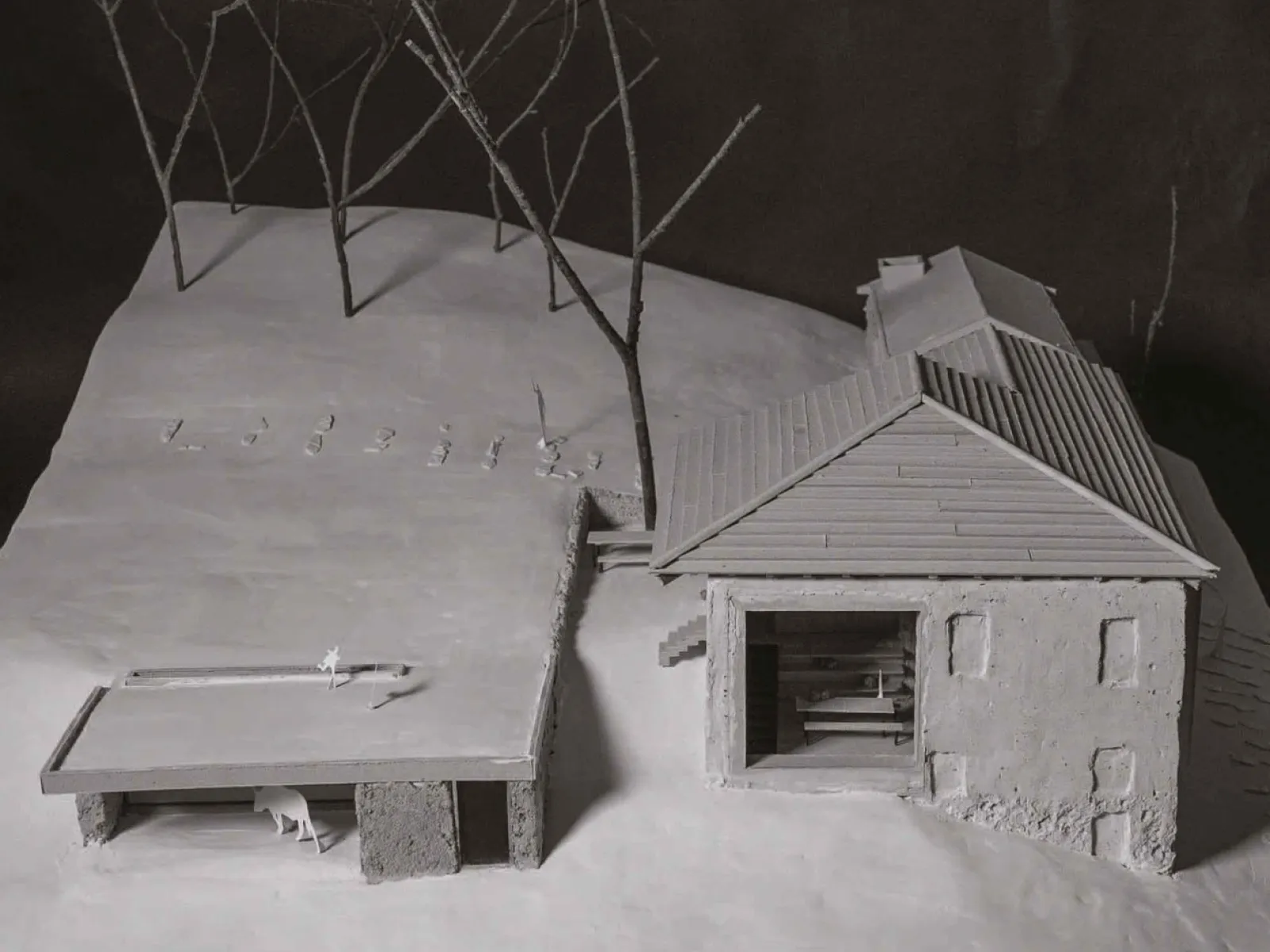 Ein Architekturmodell zeigt ein Gebäude mit Satteldach und angrenzendem offenen Unterstand auf einer schneebedeckten Landschaft. Im Hintergrund stehen stilisierte Bäume.