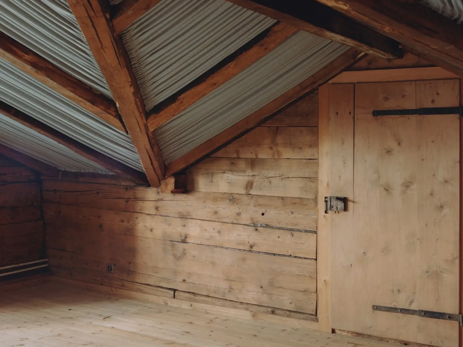 Innenansicht eines Dachbodens mit Holzwänden und Holzboden, sichtbare Dachbalken und Wellblechverkleidung. Eine Holztür befindet sich an der rechten Wand.