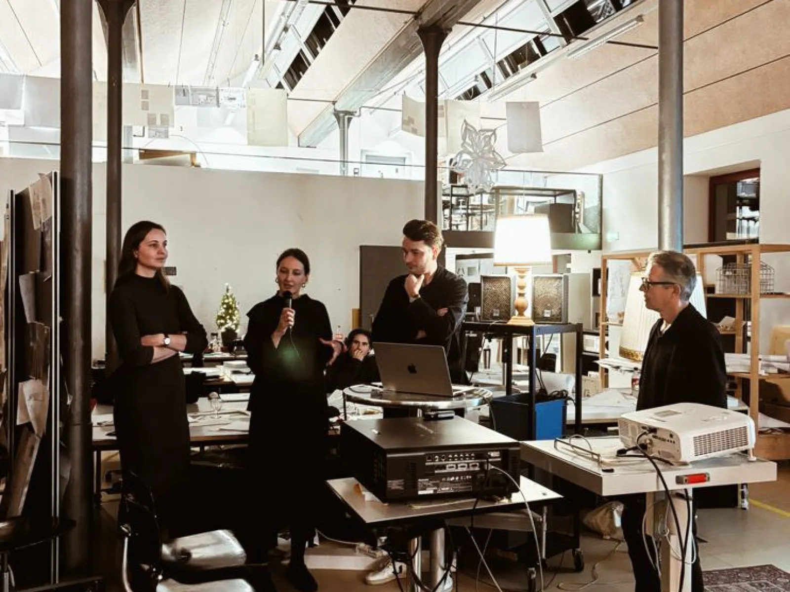 Eine Gruppe von Personen steht in einem modernen Architekturstudio. Eine Person spricht in ein Mikrofon, während andere aufmerksam zuhören. Im Raum sind technische Geräte und Präsentationsmaterialien zu sehen.