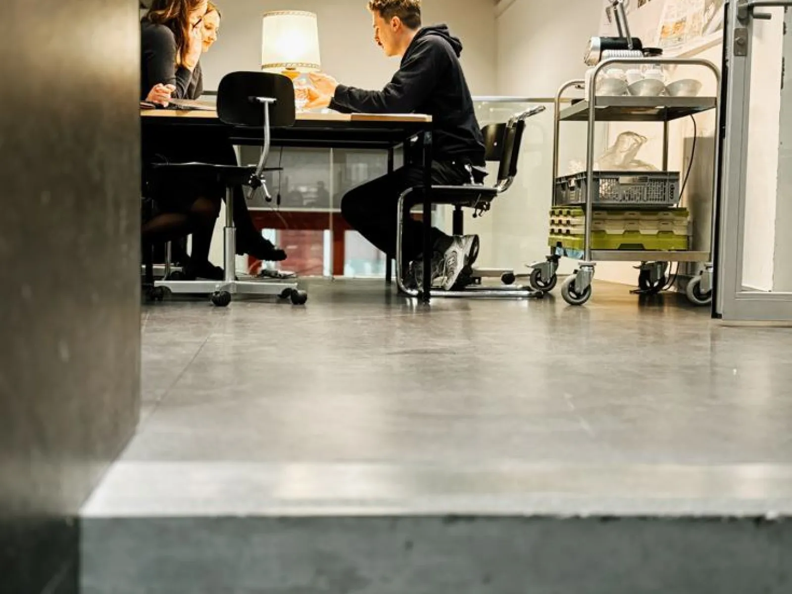 Drei Personen sitzen an einem Tisch in einem modernen Seminarraum und diskutieren. Im Hintergrund sind Architekturpläne an der Wand sichtbar. Der Raum ist hell beleuchtet und wirkt einladend.