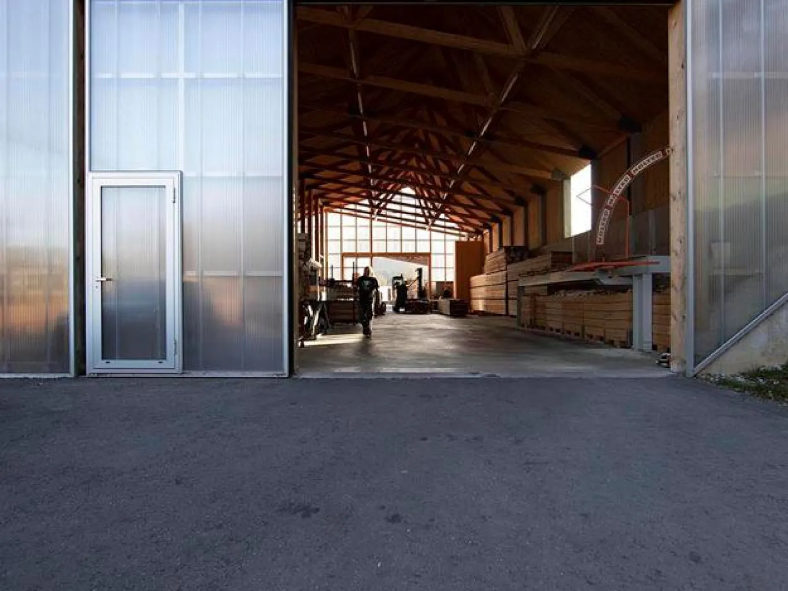Ein modernes Gebäude mit einer großen offenen Tür, die den Blick auf eine helle, hölzerne Innenstruktur freigibt. Im Inneren sind Personen und gestapelte Holzmaterialien sichtbar. Die Architektur betont klare Linien und natürliche Materialien.