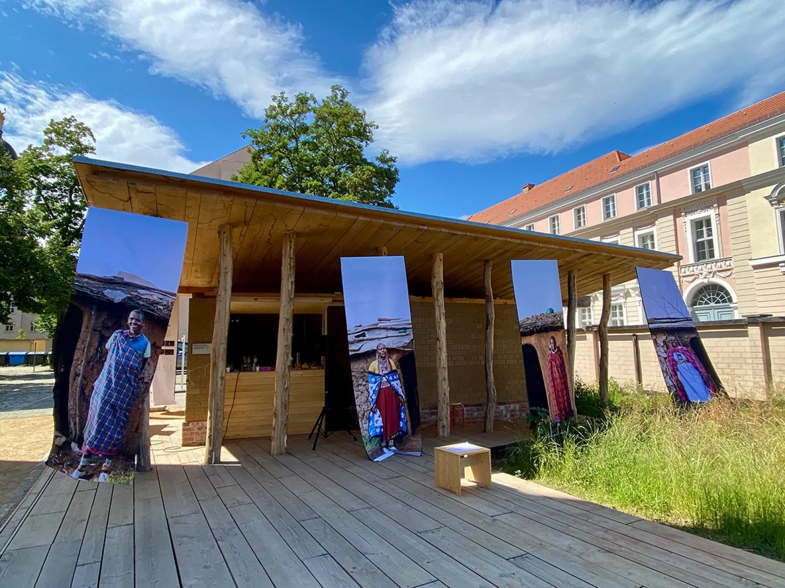 Ein Holzpavillon mit großen, vertikalen Fotobannern, die Maasai-Baumeisterinnen zeigen, steht auf einer Holzplattform. Im Hintergrund sind historische Gebäude und ein blauer Himmel mit Wolken zu sehen. Die Szene ist Teil einer Ausstellung über weibliche Perspektiven in der Architektur.
