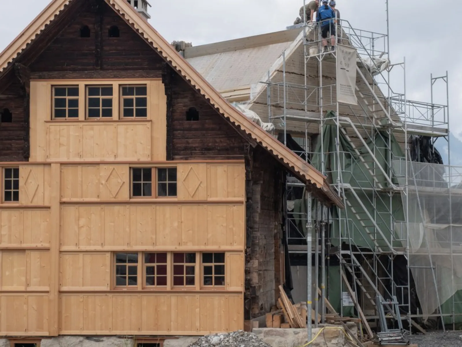 Ein traditionelles Gebäude wird renoviert. Links ist eine Holzfassade zu sehen, rechts stehen Gerüste mit Bauarbeitern auf dem Dach. Der Himmel ist bewölkt.