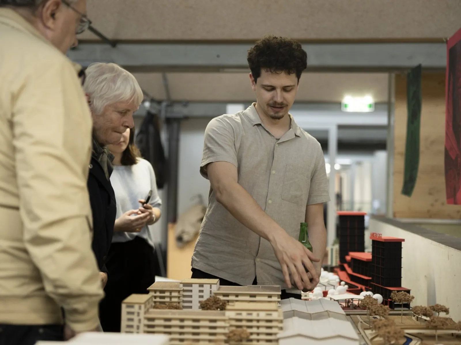 Eine Gruppe von Menschen betrachtet ein Architekturmodell in einem Innenraum. Ein Mann zeigt auf das Modell und erklärt es den anderen Anwesenden. Im Hintergrund sind weitere Modelle und Regale zu sehen.