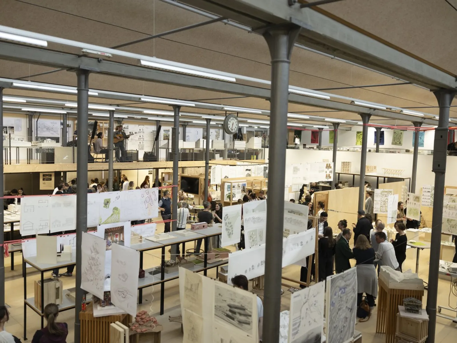 Innenansicht eines belebten Architekturstudios mit zahlreichen Menschen, die sich unterhalten und Arbeiten betrachten. Zeichnungen und Pläne hängen von der Decke. Die Szene vermittelt eine kreative und kollaborative Atmosphäre.