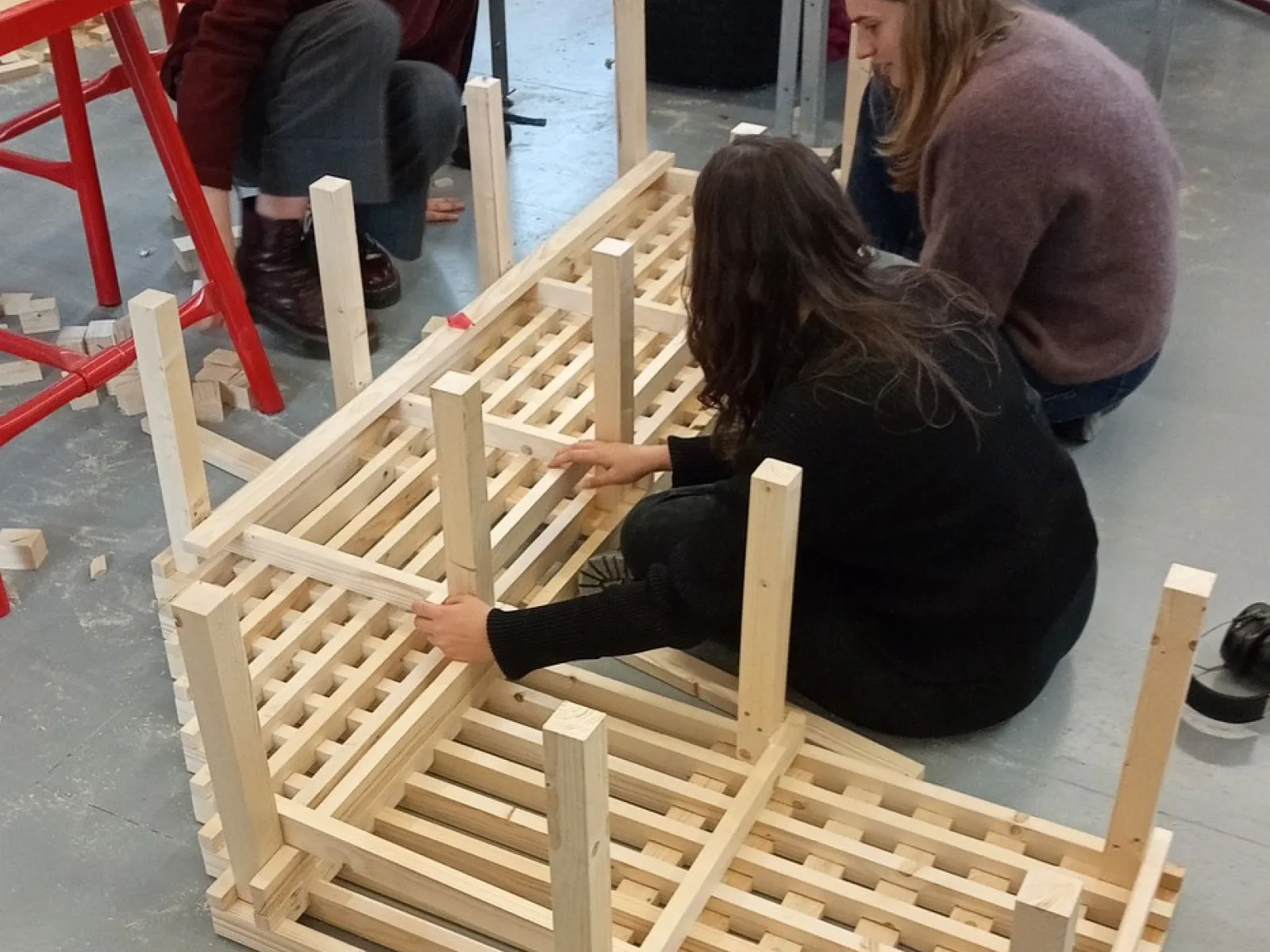 Drei Studierende arbeiten gemeinsam an einem Holzprojekt in einer Werkstatt. Sie montieren ein Möbelstück aus Holzlatten. Im Hintergrund sind Werkbänke und Werkzeuge sichtbar.
