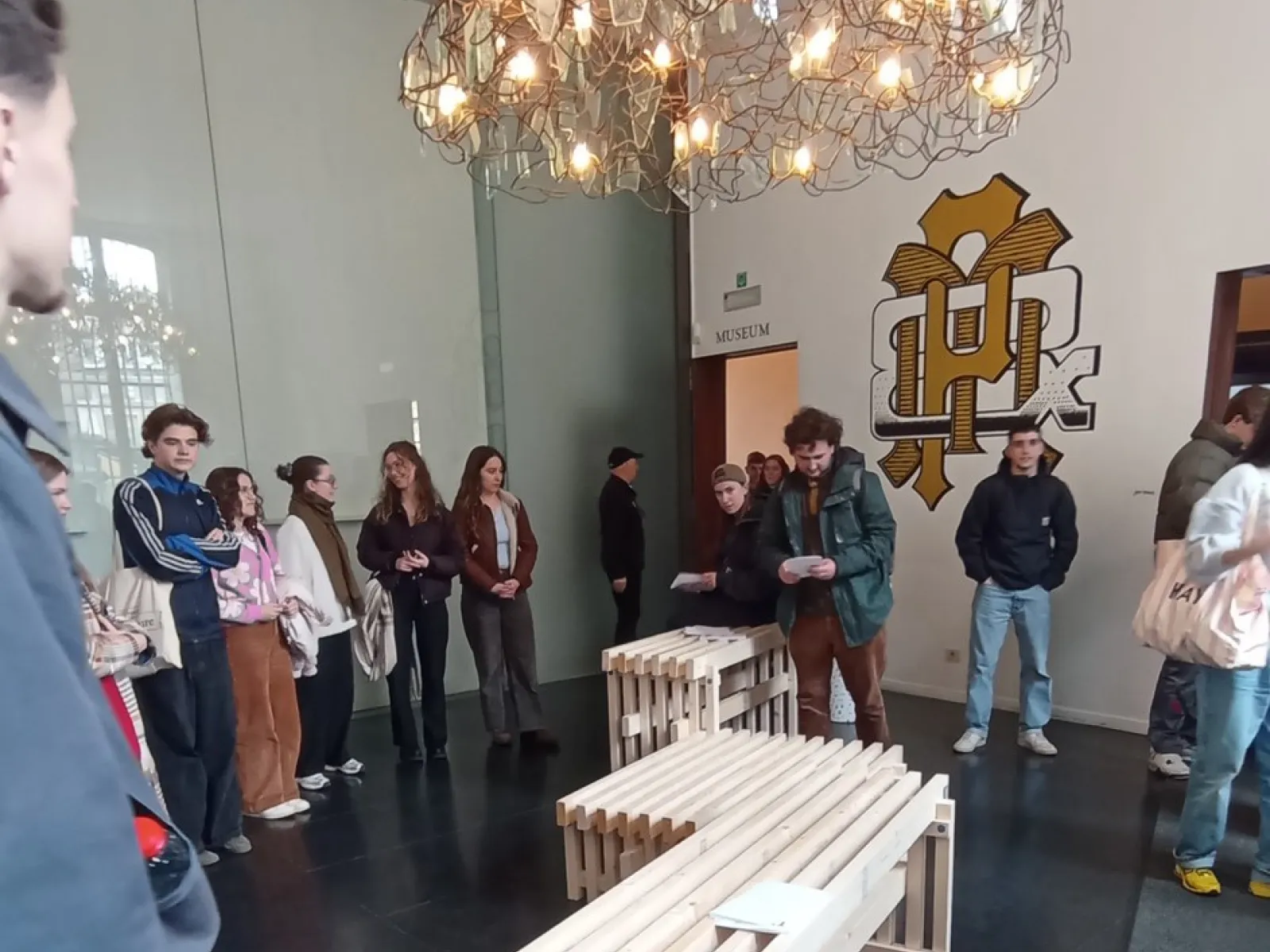 Eine Gruppe von Studierenden steht in einem Raum der Universität Antwerpen um ein modernes, aus Holzlatten gefertigtes Möbelstück. Über ihnen hängt eine große, kunstvolle Lampe. Im Hintergrund ist ein Wandlogo der Universität zu sehen.