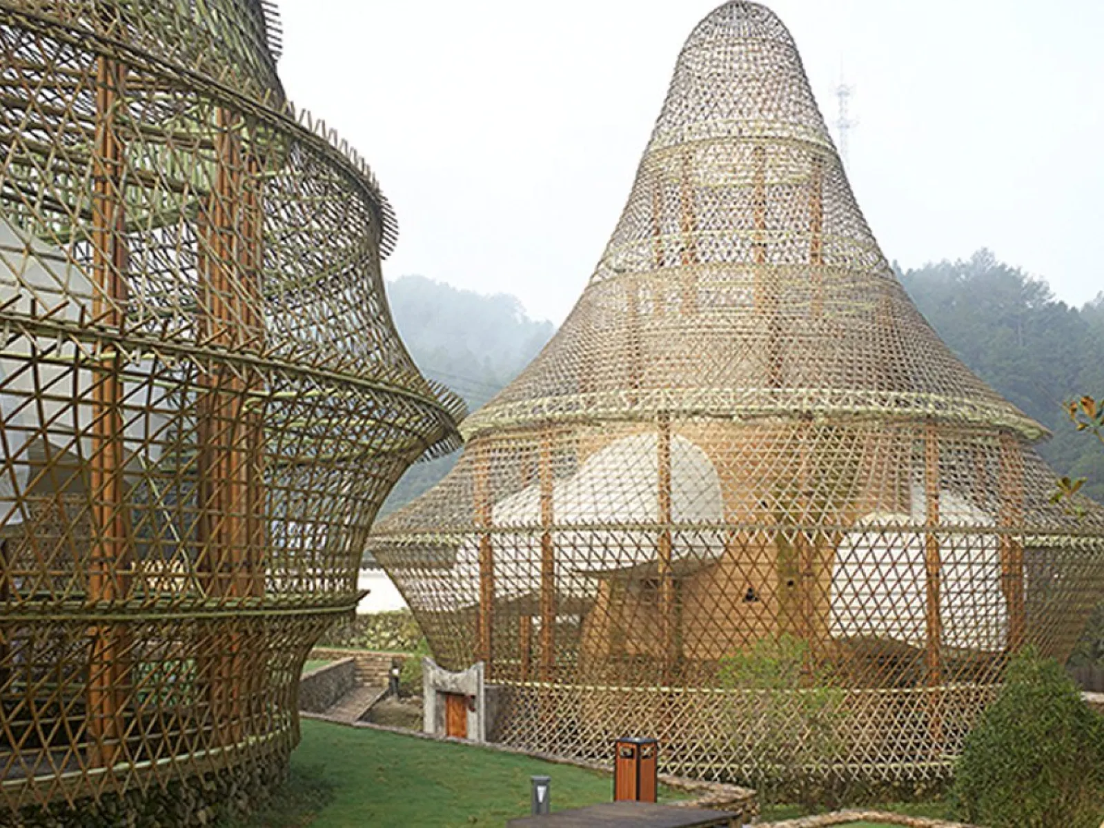 Bambusarchitektur mit kuppelförmigen Strukturen in einer grünen Landschaft. Die Konstruktionen zeigen innovative Nutzung natürlicher Materialien und verbinden traditionelle Bauweisen mit modernem Design.