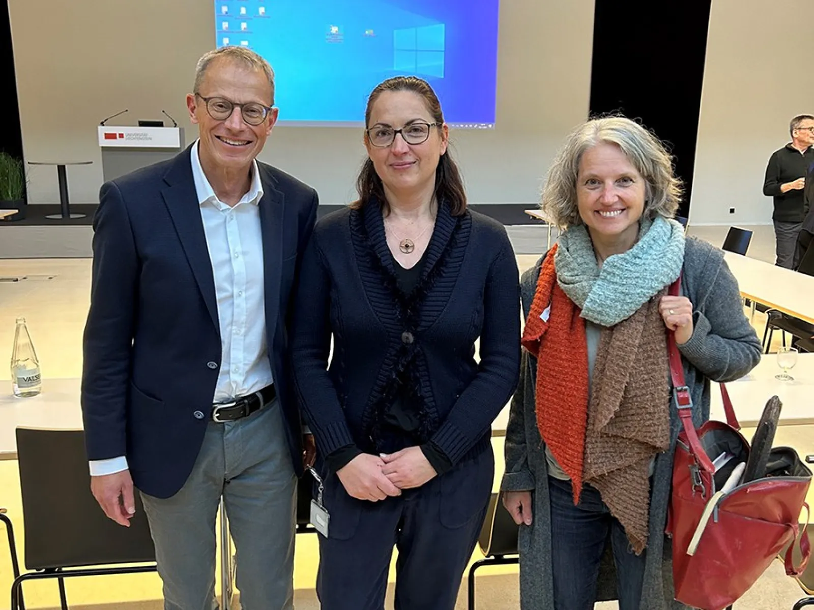 Drei Personen stehen lächelnd in einem Auditorium der Universität Liechtenstein. Im Hintergrund sind ein Rednerpult und eine Leinwand mit einer Präsentation zu sehen.