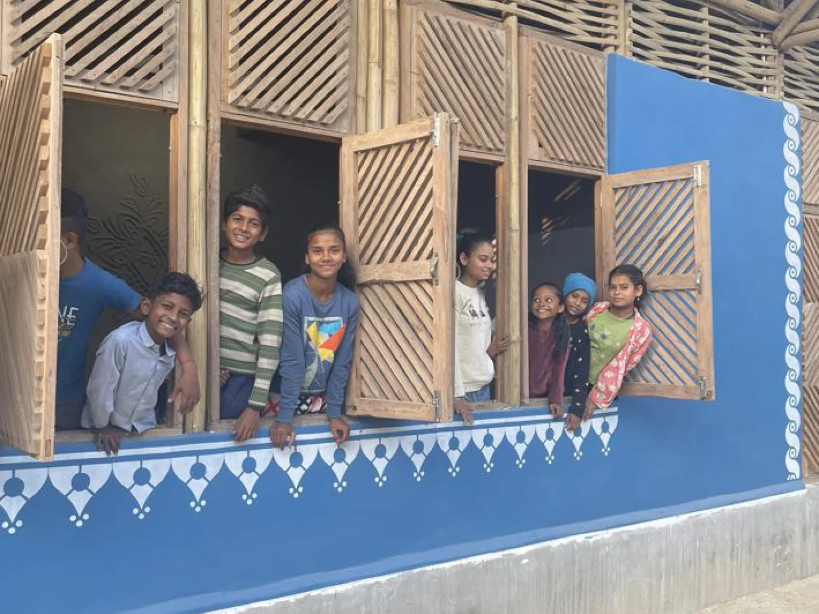 Kinder schauen lächelnd aus den Fenstern eines neu gebauten Schulgebäudes in Bihar, Indien. Die Fassade ist blau mit dekorativen weißen Mustern.