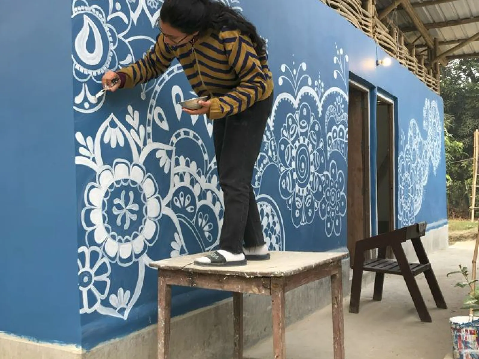 Eine Person bemalt die blaue Außenwand einer Schule in Indien mit weißen, floralen Mustern. Sie steht auf einem Holztisch, um die oberen Bereiche zu erreichen. Das Gebäude hat ein Wellblechdach und ist Teil eines Bildungsprojekts.