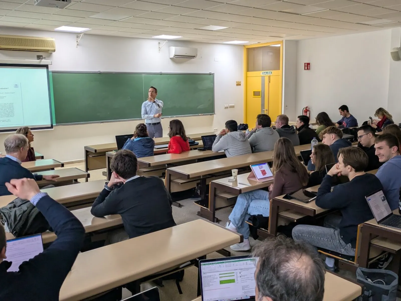 Ein Dozent hält einen Vortrag in einem Hörsaal, umgeben von Studierenden, die aufmerksam zuhören und Notizen auf Laptops machen. Im Hintergrund ist eine Tafel und eine Präsentation zu sehen.