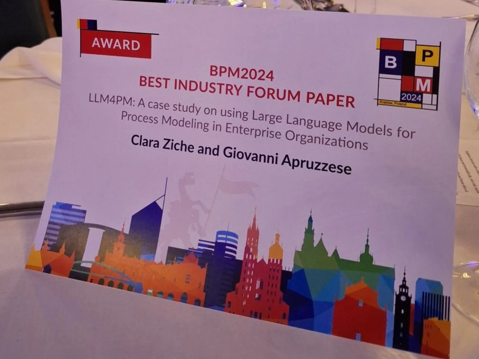 Ein Tischaufsteller mit der Aufschrift "BPM2024 Best Industry Forum Paper" und dem Titel der ausgezeichneten Masterarbeit von Clara Ziche und Giovanni Apruzzese. Im Hintergrund sind stilisierte Skyline-Elemente zu sehen.
