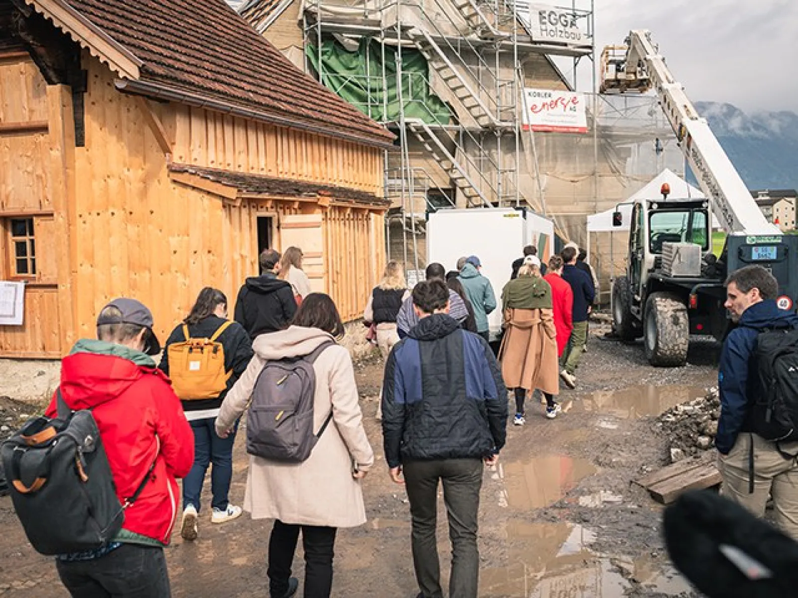 Eine Gruppe von Menschen besichtigt eine Baustelle mit einem Holzgebäude und Baugerüsten. Im Hintergrund sind Berge zu sehen. Die Szene wirkt lebendig und zeigt das Interesse an nachhaltigem Bauen.