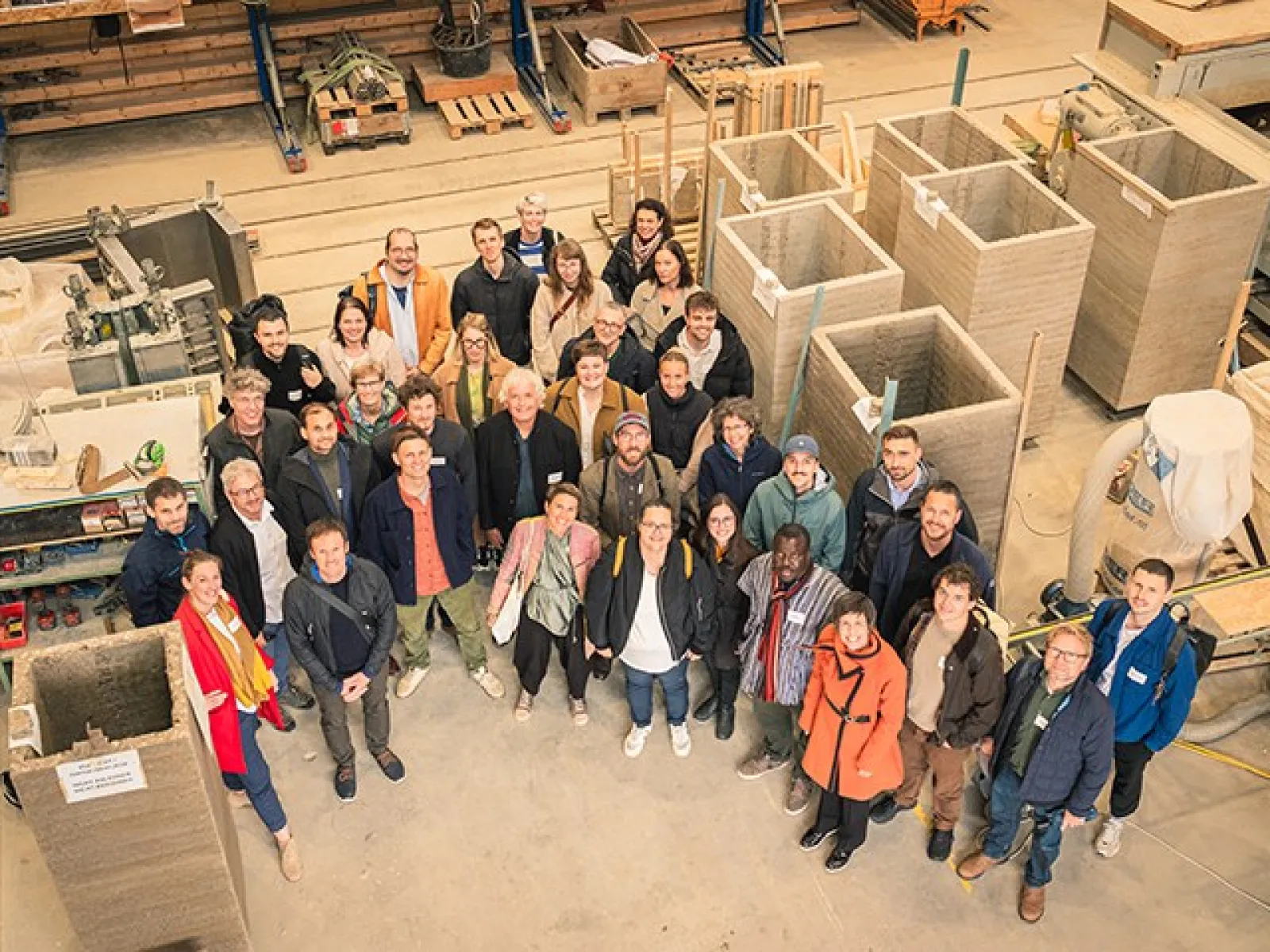 Gruppenfoto von Teilnehmern des Earth-Hub-Symposiums in einer Werkstatt, umgeben von Baumaterialien wie Stampflehm-Elementen.