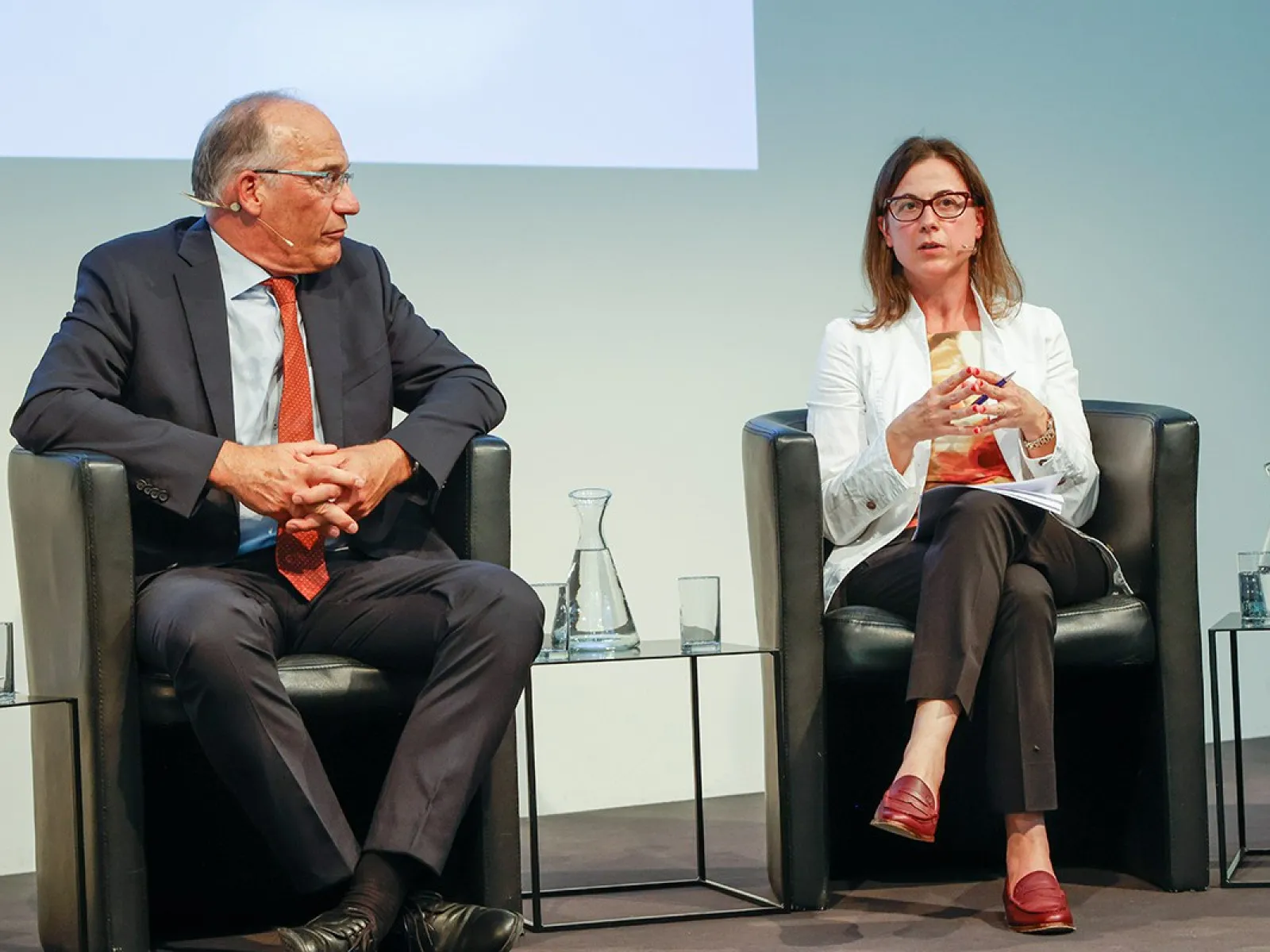 Zwei Personen sitzen auf einer Bühne in schwarzen Sesseln. Die Person links trägt einen dunklen Anzug und eine rote Krawatte, die Person rechts trägt eine weiße Jacke und rote Schuhe. Zwischen ihnen stehen Wasserkaraffen und Gläser.