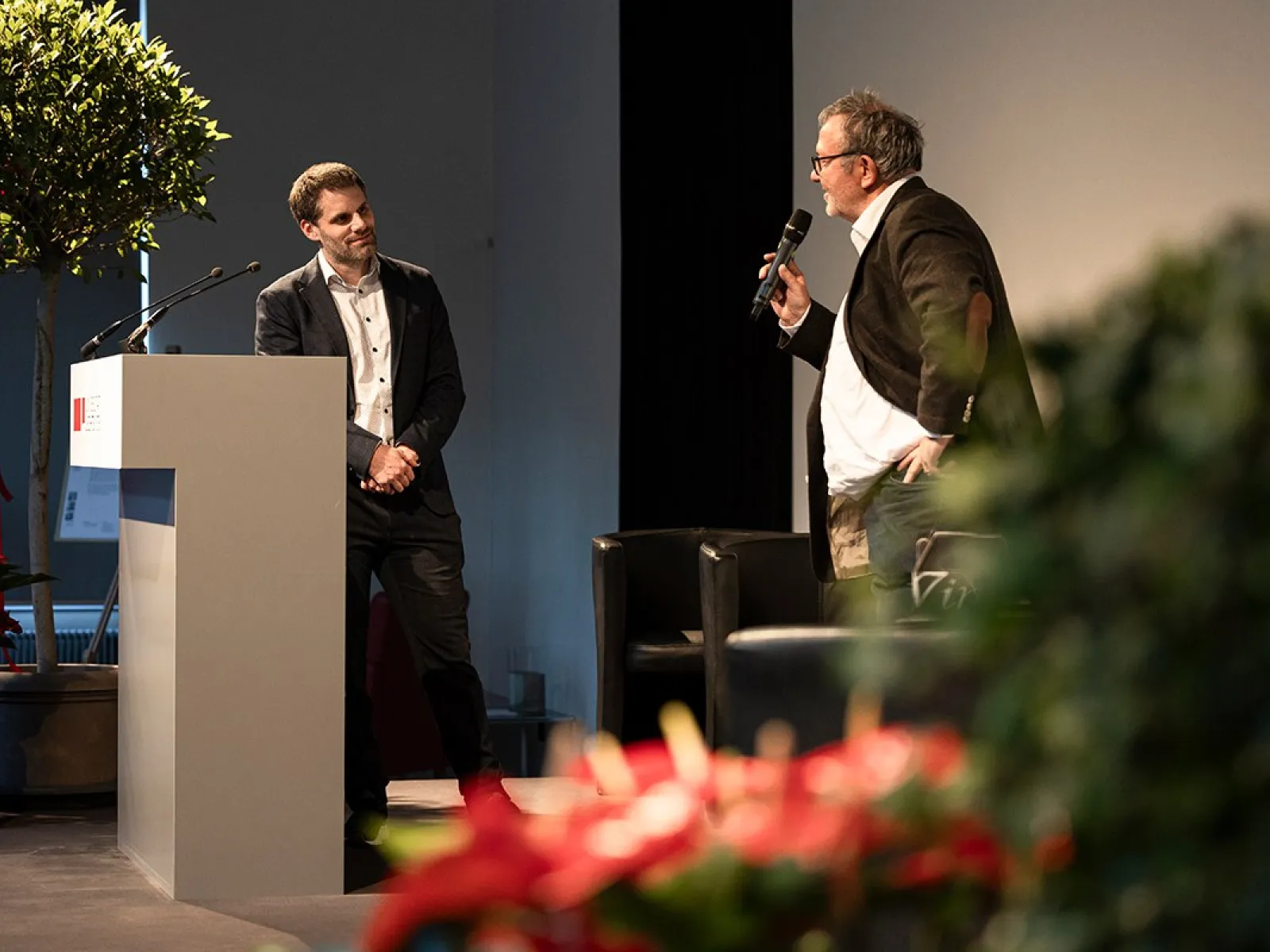 Zwei Männer stehen auf einer Bühne bei einer Veranstaltung. Einer spricht in ein Mikrofon, während der andere an einem Rednerpult steht. Im Hintergrund sind Pflanzen und ein großer Bildschirm zu sehen.