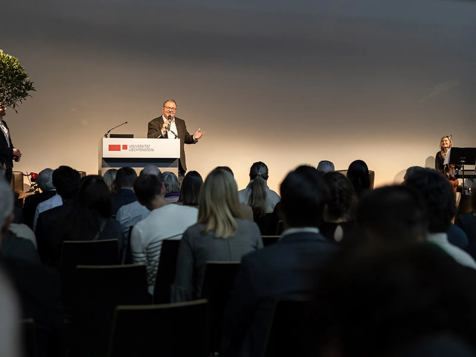 Ein Redner steht an einem Podium mit dem Logo der Universität Liechtenstein, spricht zu einem Publikum. Im Hintergrund sind zwei Musiker zu sehen. Die Veranstaltung findet in einem großen Saal statt.