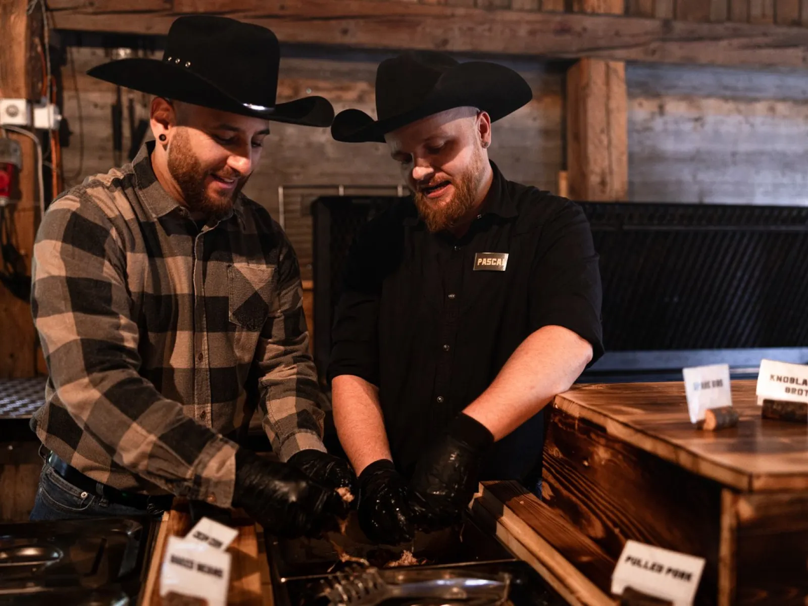 Zwei Personen in Cowboyhüten bereiten Essen an einem Buffet vor. Sie tragen schwarze Handschuhe und stehen in einem rustikalen Raum mit Holzwänden. Vor ihnen sind Schilder mit verschiedenen Fleischsorten zu sehen.