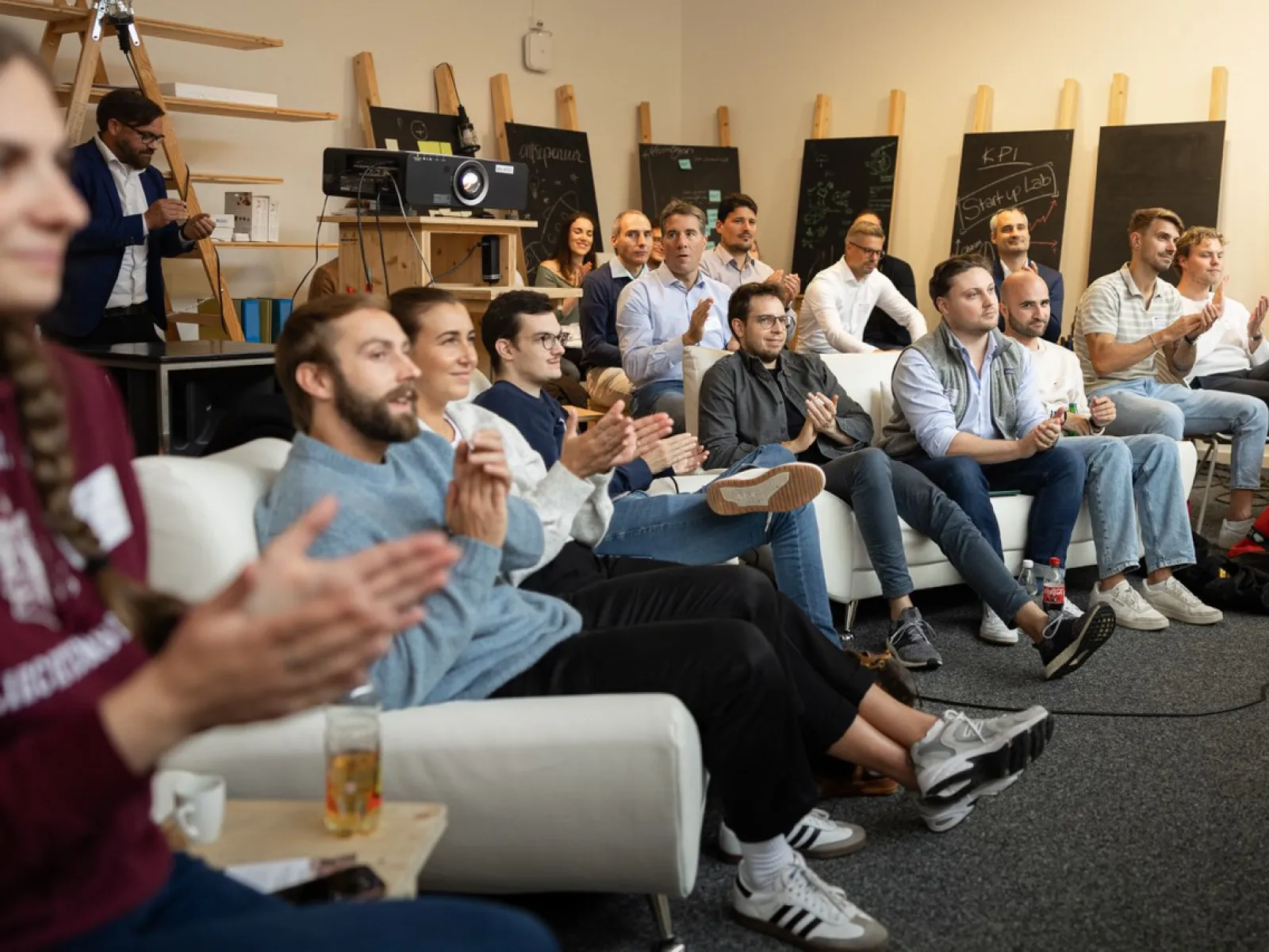 Eine Gruppe von Menschen sitzt in einem Raum und applaudiert. Im Hintergrund sind Tafeln und technische Geräte zu sehen. Die Atmosphäre wirkt konzentriert und feierlich.