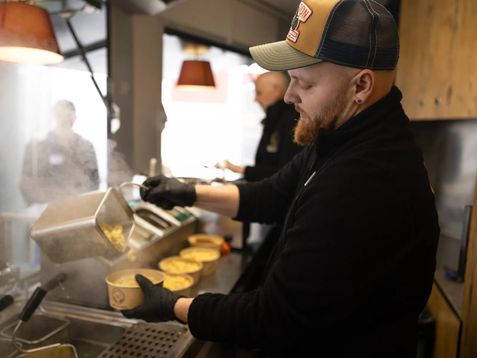 Ein Koch füllt dampfende Nudeln in Schalen in einem Foodtruck. Im Hintergrund sind weitere Personen und Küchenutensilien zu sehen.