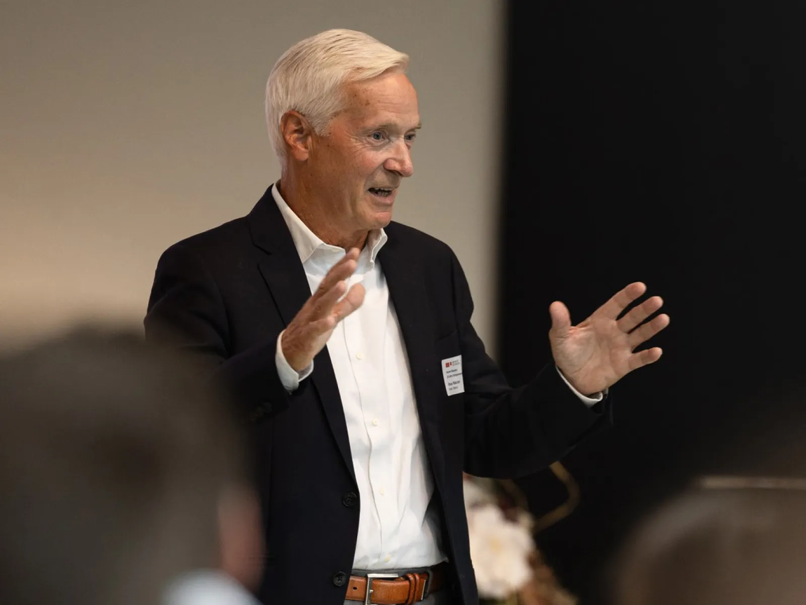 Ein älterer Mann in einem dunklen Anzug spricht gestikulierend bei einer Veranstaltung. Im Hintergrund sind unscharfe Personen zu erkennen.