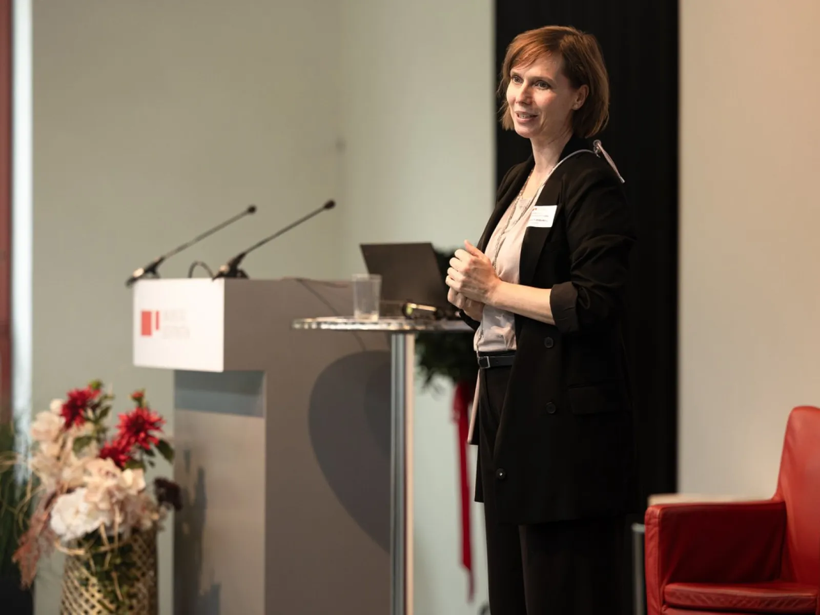 Eine Person steht auf einer Bühne neben einem Rednerpult mit Mikrofonen. Im Hintergrund sind Blumenarrangements und ein roter Stuhl zu sehen.