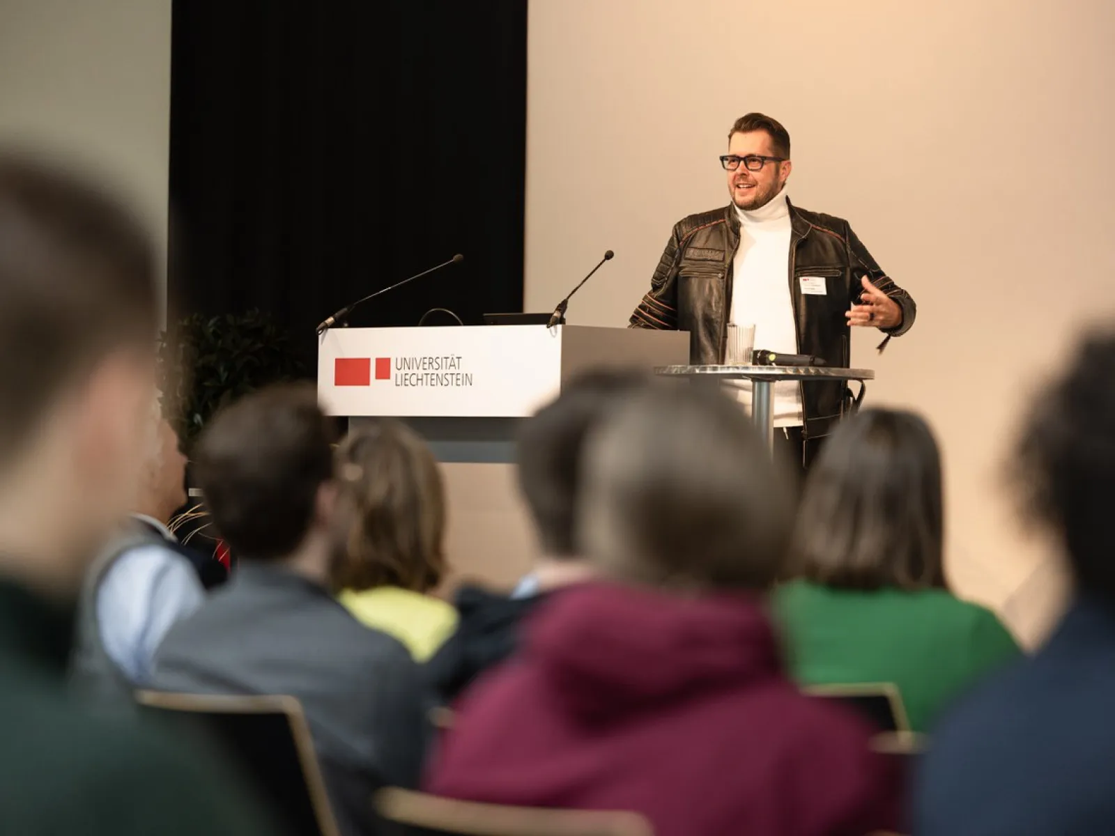 Ein Redner mit schwarzer Lederjacke steht auf der Bühne vor dem Redepult und hält eine Rede. Im vorderen Teil des Bildes ist ein Teil des Publikums zu sehen.