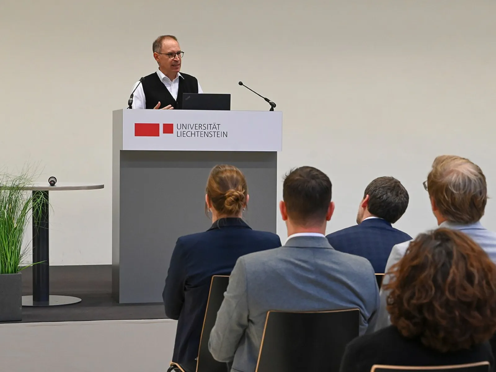 Ein Redner steht an einem Podium mit dem Logo der Universität Liechtenstein und spricht zu einem sitzenden Publikum. Im Hintergrund sind Pflanzen zu sehen.