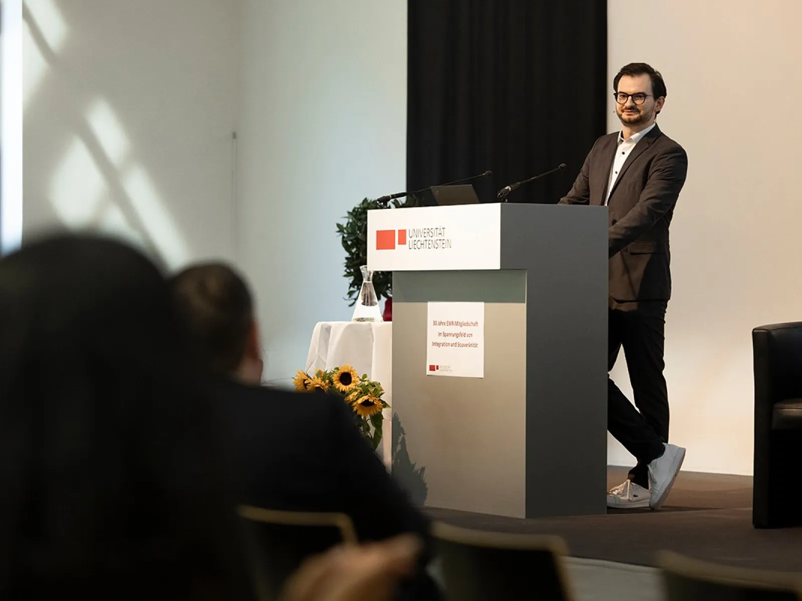 Ein Mann steht an einem Rednerpult in einem Konferenzraum der Universität Liechtenstein. Er trägt einen Anzug und spricht zu einem Publikum. Im Hintergrund sind Sonnenblumen und ein Fenster zu sehen.