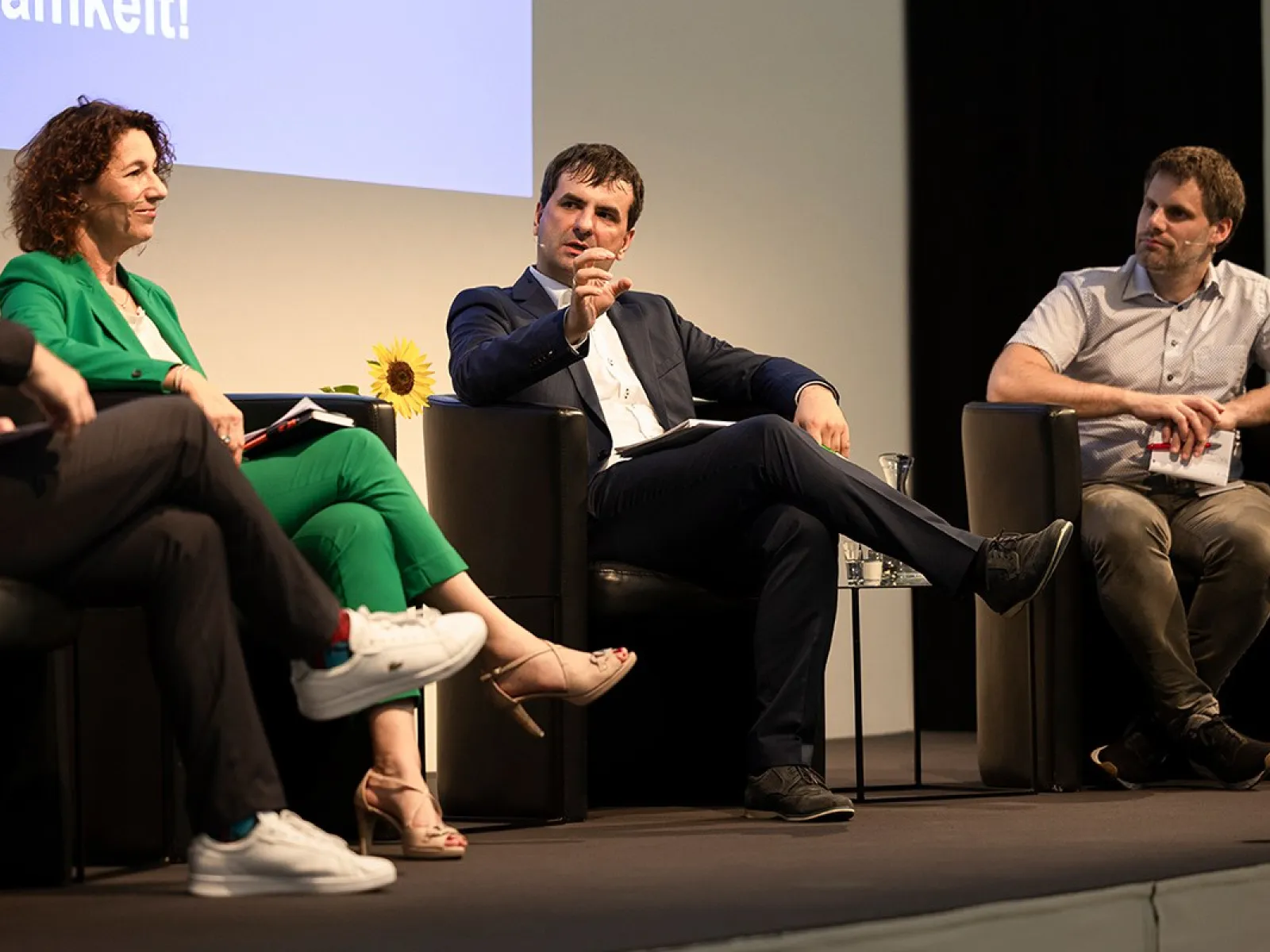Podiumsdiskussion an der Universität Liechtenstein: Drei Personen sitzen auf einer Bühne in schwarzen Sesseln und diskutieren angeregt. Eine Frau in grünem Anzug und zwei Männer in formeller Kleidung sind zu sehen. Im Hintergrund ist ein Teil einer Leinwand mit Text sichtbar.