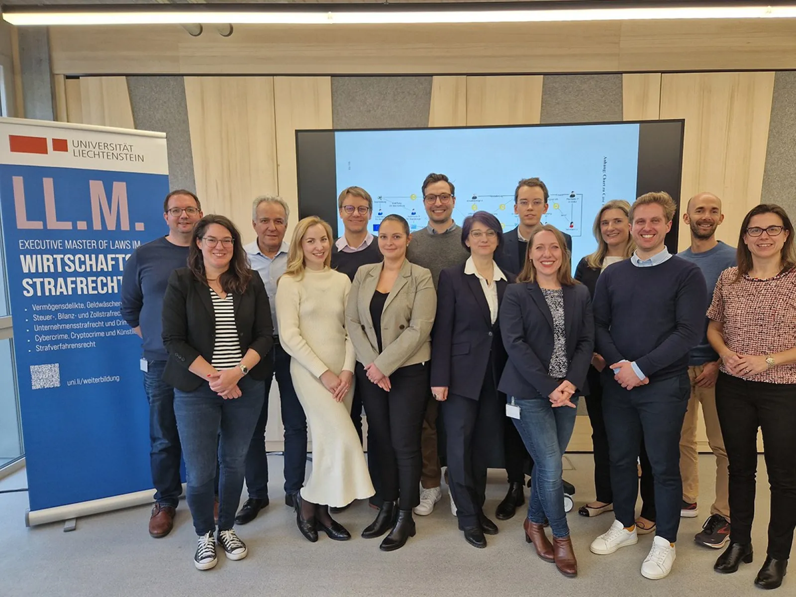 Gruppenfoto von Studierenden und Dozenten des Executive Masters of Law im Wirtschaftsstrafrecht an der Universität Liechtenstein. Im Hintergrund ein Bildschirm mit Präsentationsfolie und ein Roll-up mit Informationen zum Studiengang.