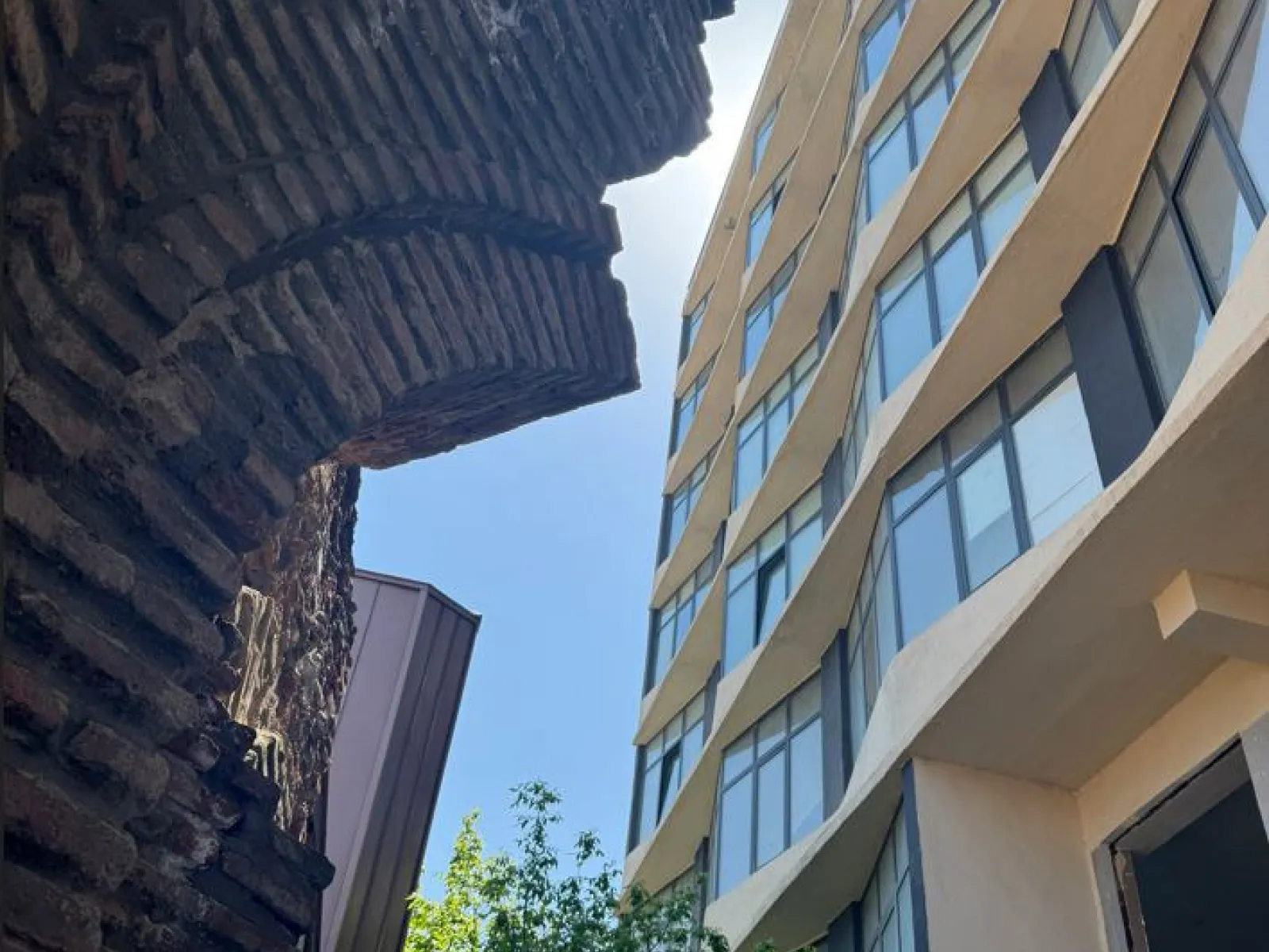 Historische Backsteinmauer im Kontrast zu einem modernen, wellenförmigen Glasgebäude bei Tageslicht.