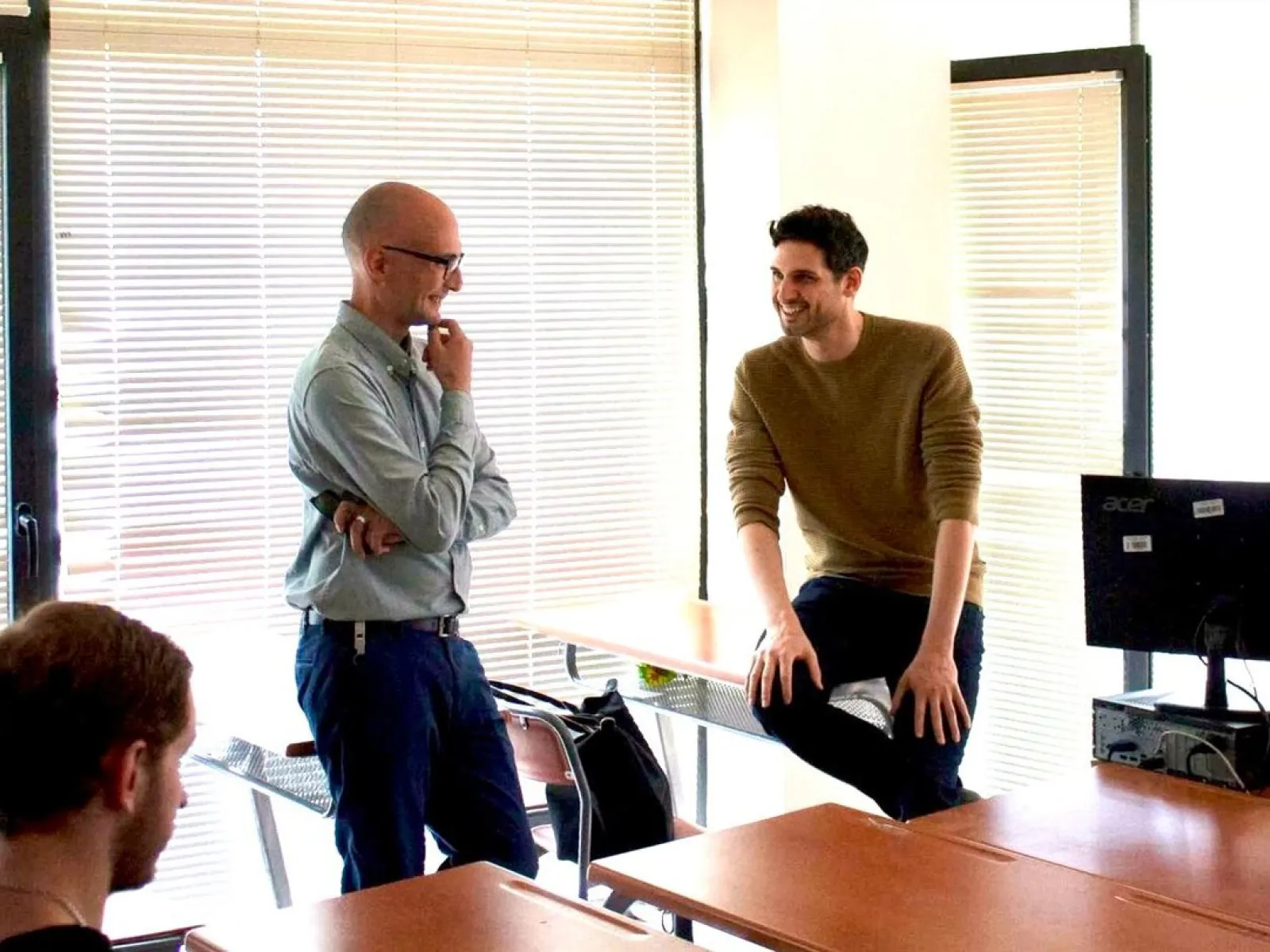Zwei Männer in einem hellen Raum stehen und sitzen lächelnd an einem Tisch, in einem Gespräch vertieft. Im Hintergrund sind Jalousien zu sehen.