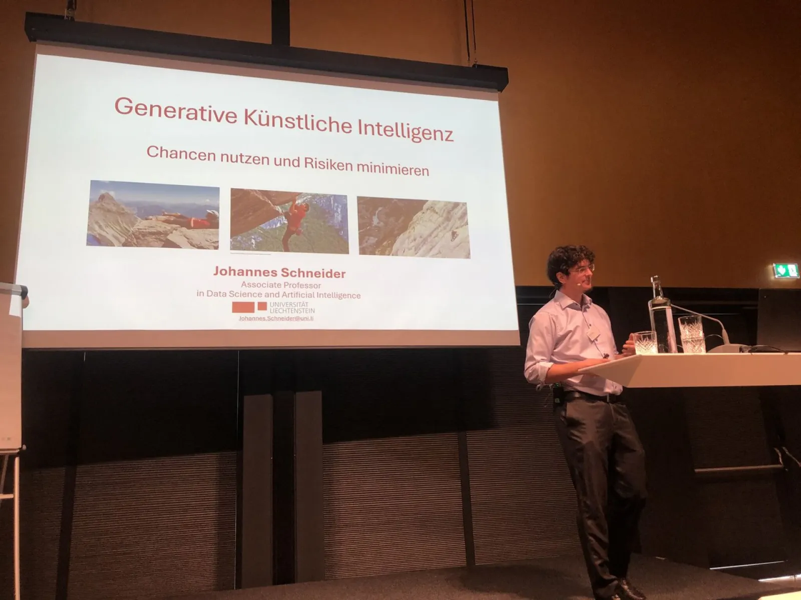 Vortragender steht vor einer Präsentation mit dem Titel "Generative Künstliche Intelligenz: Chancen nutzen und Risiken minimieren". Der Raum ist dunkel, der Fokus liegt auf der Leinwand und dem Redner.