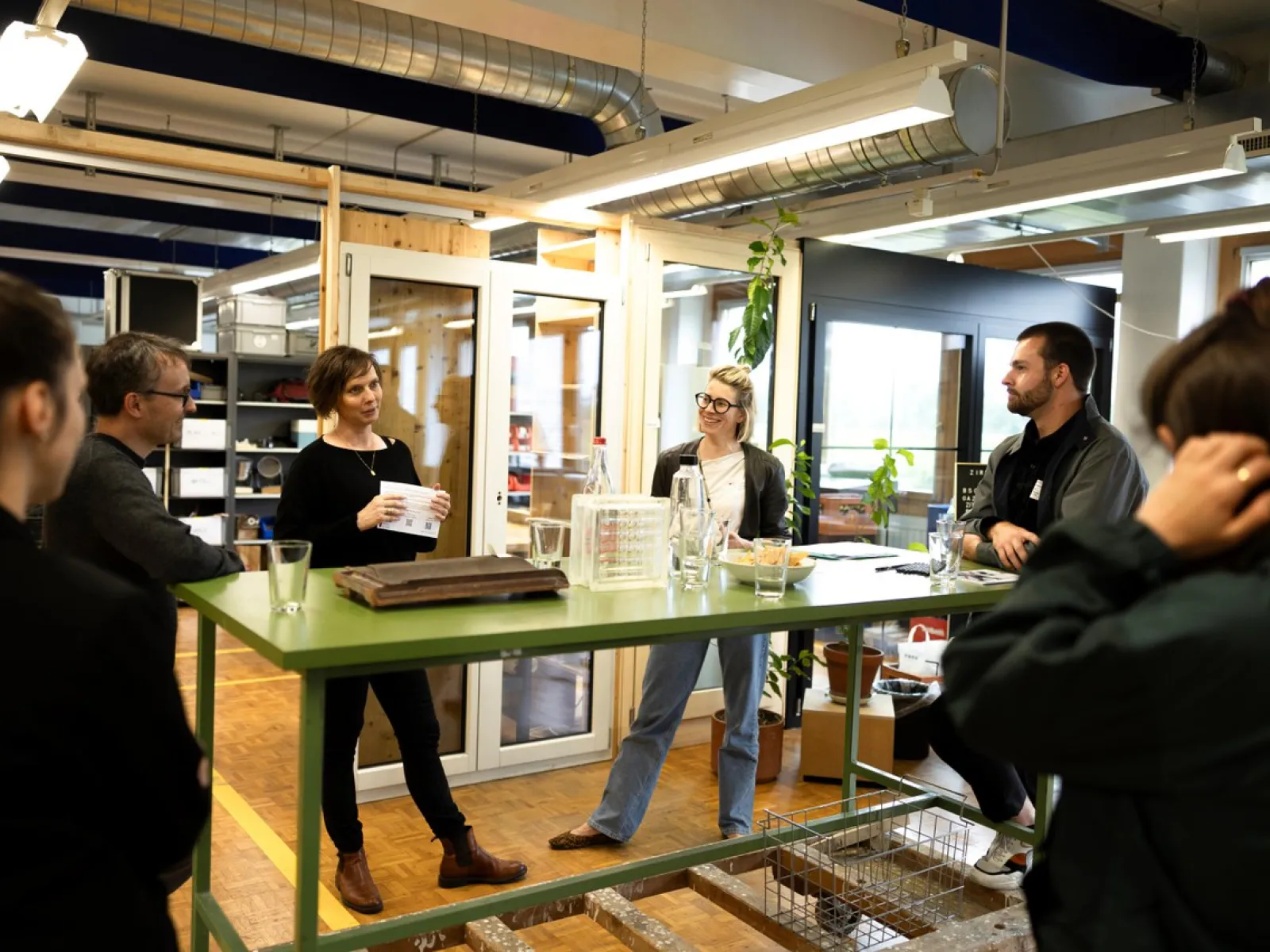 Personen stehen um einen grünen Tisch in einem modernen Raum mit Glaswänden und Pflanzen. Eine Person hält ein Papier und spricht zu den anderen. Die Szene wirkt wie ein informelles Treffen oder eine Diskussion.