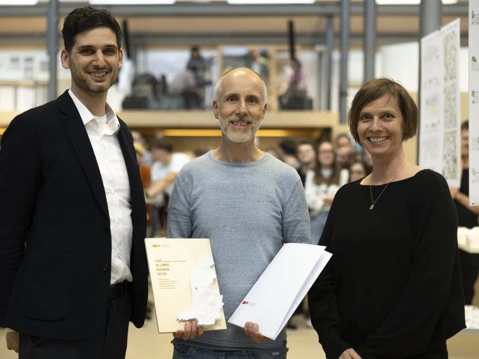 Drei Personen stehen lächelnd in einem Innenraum. Eine Person hält ein Zertifikat in der Hand. Im Hintergrund sind unscharf weitere Menschen zu sehen.