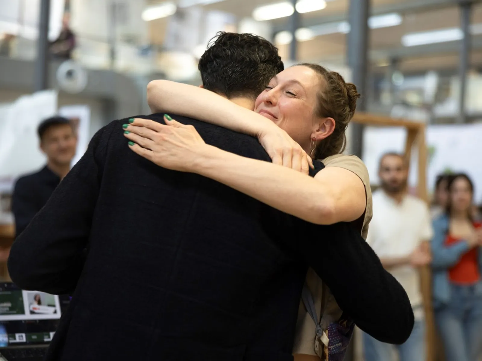 Zwei Personen umarmen sich herzlich in einem Innenraum während des LSA Sommerfests an der Universität Liechtenstein. Im Hintergrund sind unscharf weitere Personen und architektonische Details zu erkennen.