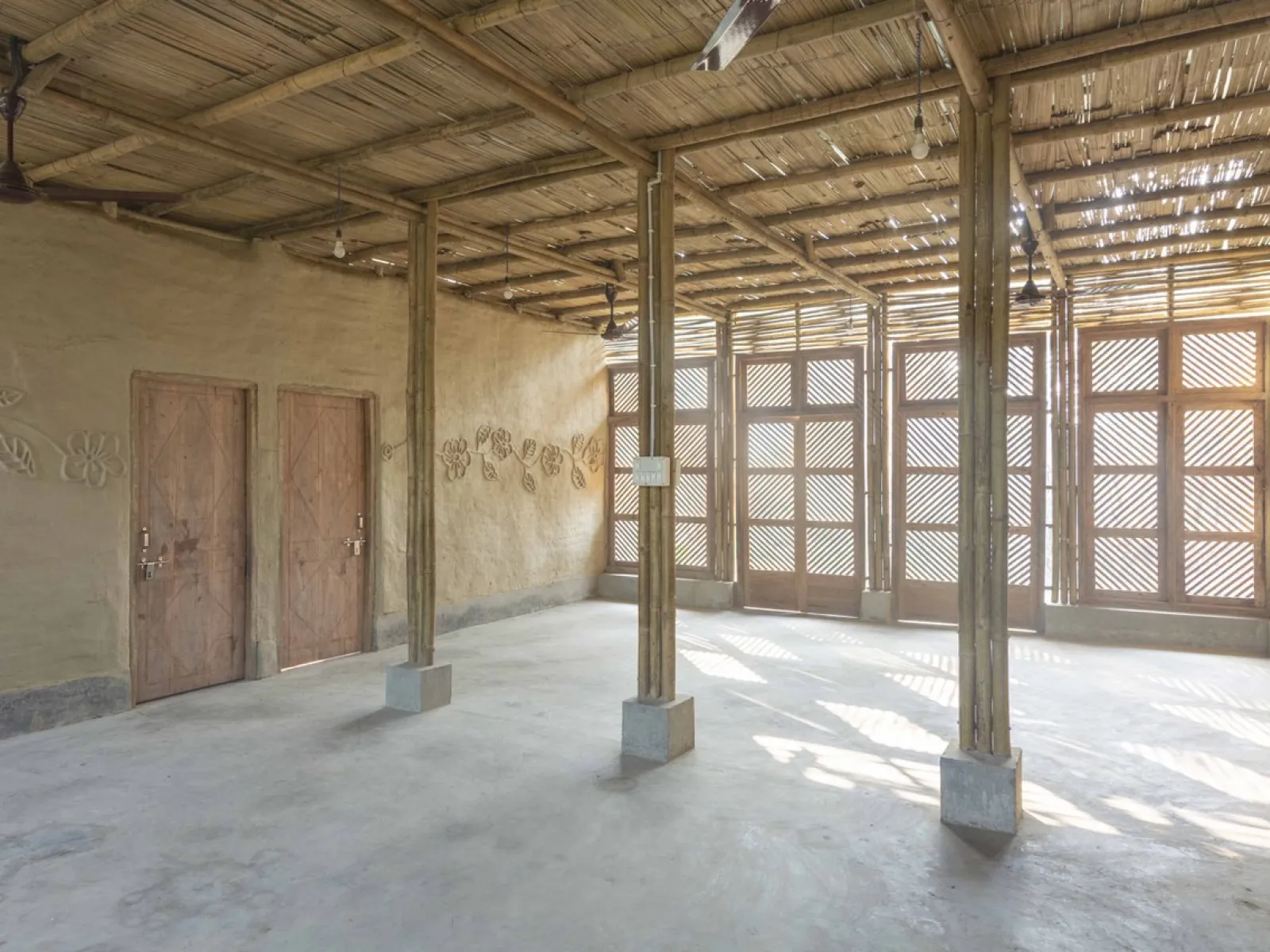 Alt-Text: Innenansicht eines nachhaltig gebauten Raumes mit Bambusstrukturen, Lehmwänden und Holztüren. Sonnenlicht fällt durch ein Gittermuster an den Fenstern und erzeugt ein warmes Ambiente.