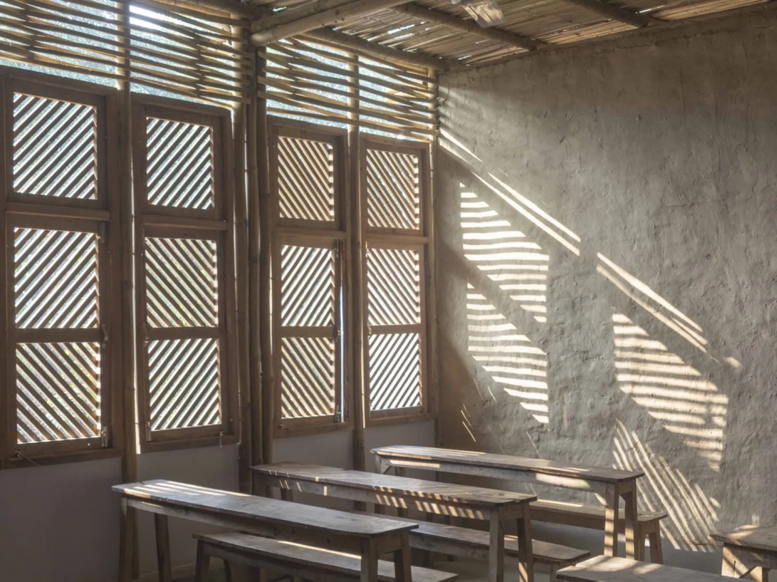 Innenraum eines nachhaltig gebauten Klassenzimmers mit Holzbänken und Wänden aus natürlichen Materialien. Sonnenlicht fällt durch hölzerne Fensterläden und erzeugt Muster auf der Wand.