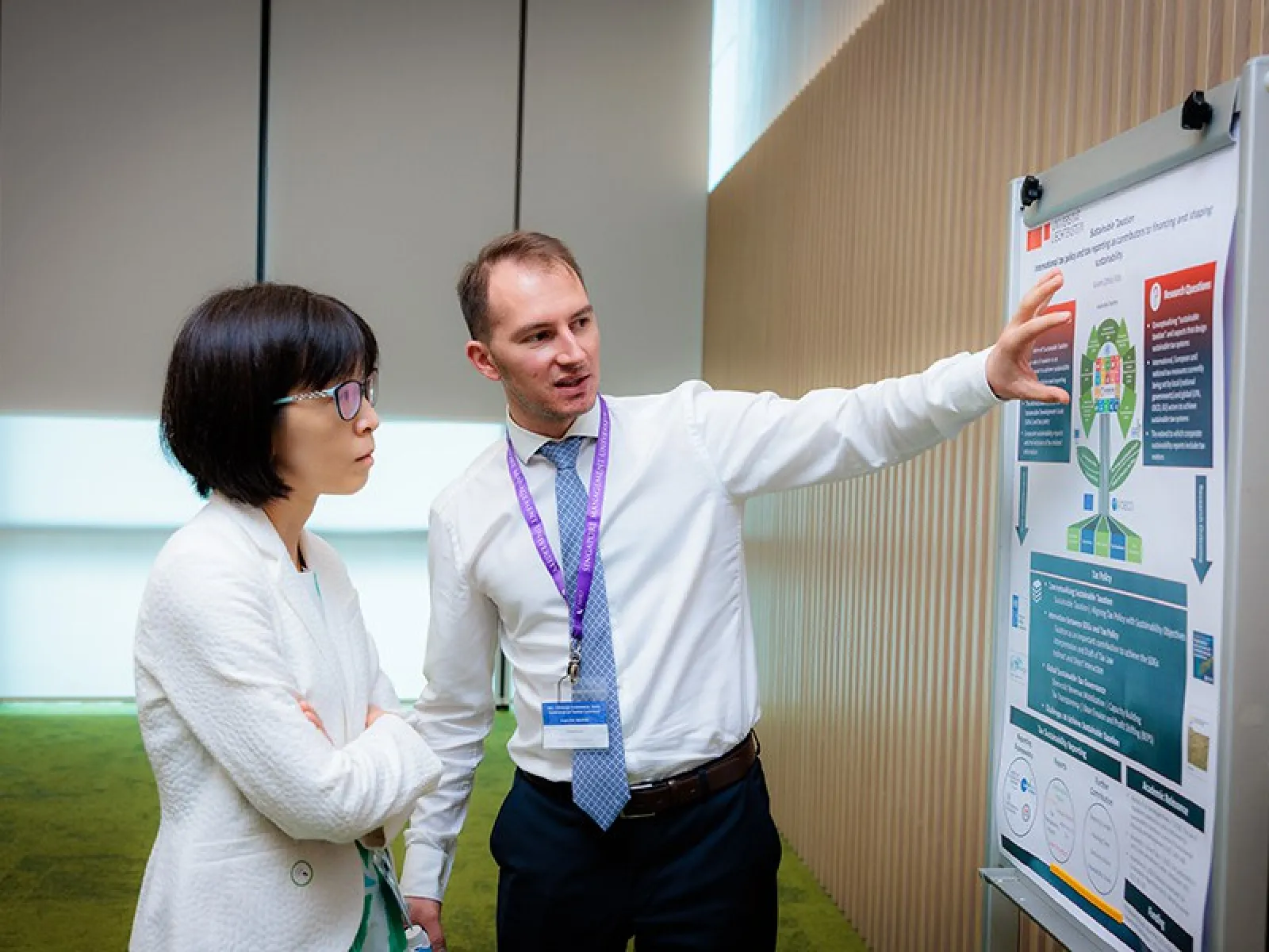 Ein Mann erklärt einer Frau ein wissenschaftliches Poster über nachhaltige Besteuerung in einem Konferenzraum.